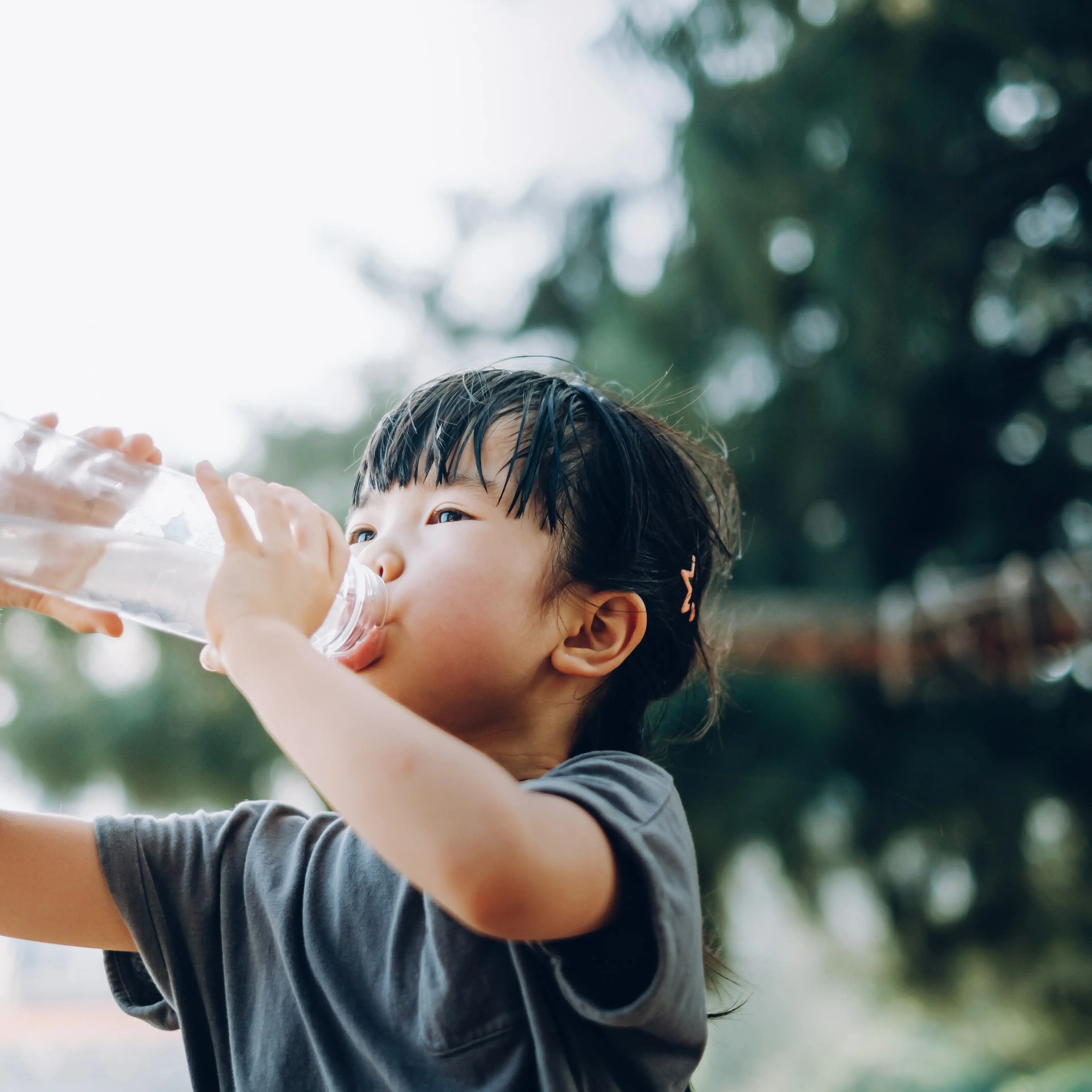 Little Asian girl is taking a big gulp of water from a reusable plastic water bottle outdoors on a sunny Summer day. Staying hydrated. Healthy lifestyle concept