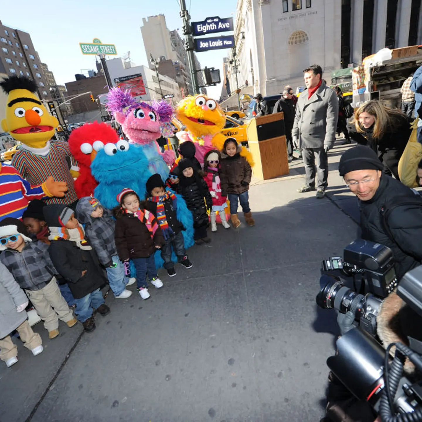 A group of children stand in front of the main characters of Sesame Street while they are photographed by a crowd of the press.