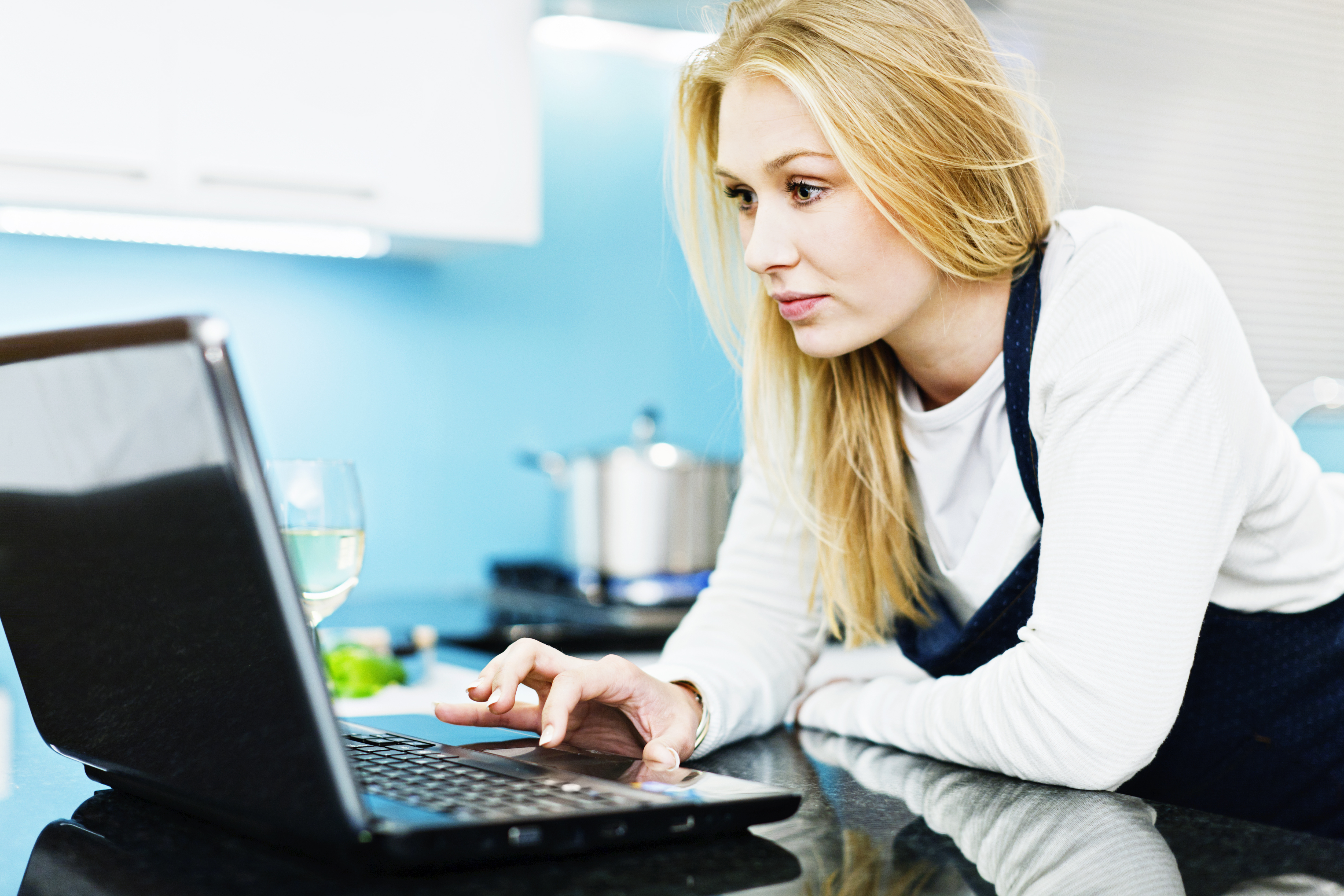 Young blonde woman in kitchen preparing food checks laptop