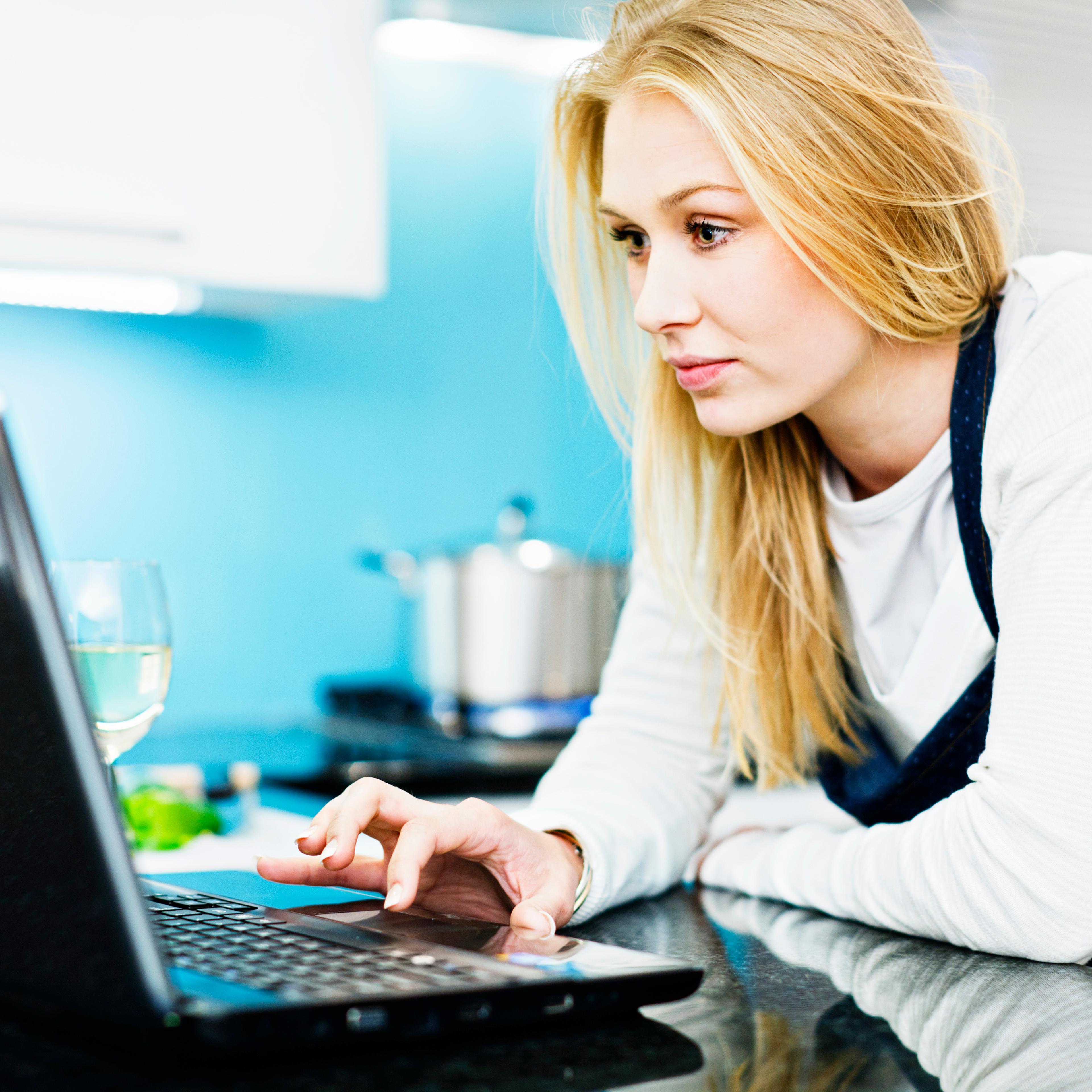 Young blonde woman in kitchen preparing food checks laptop