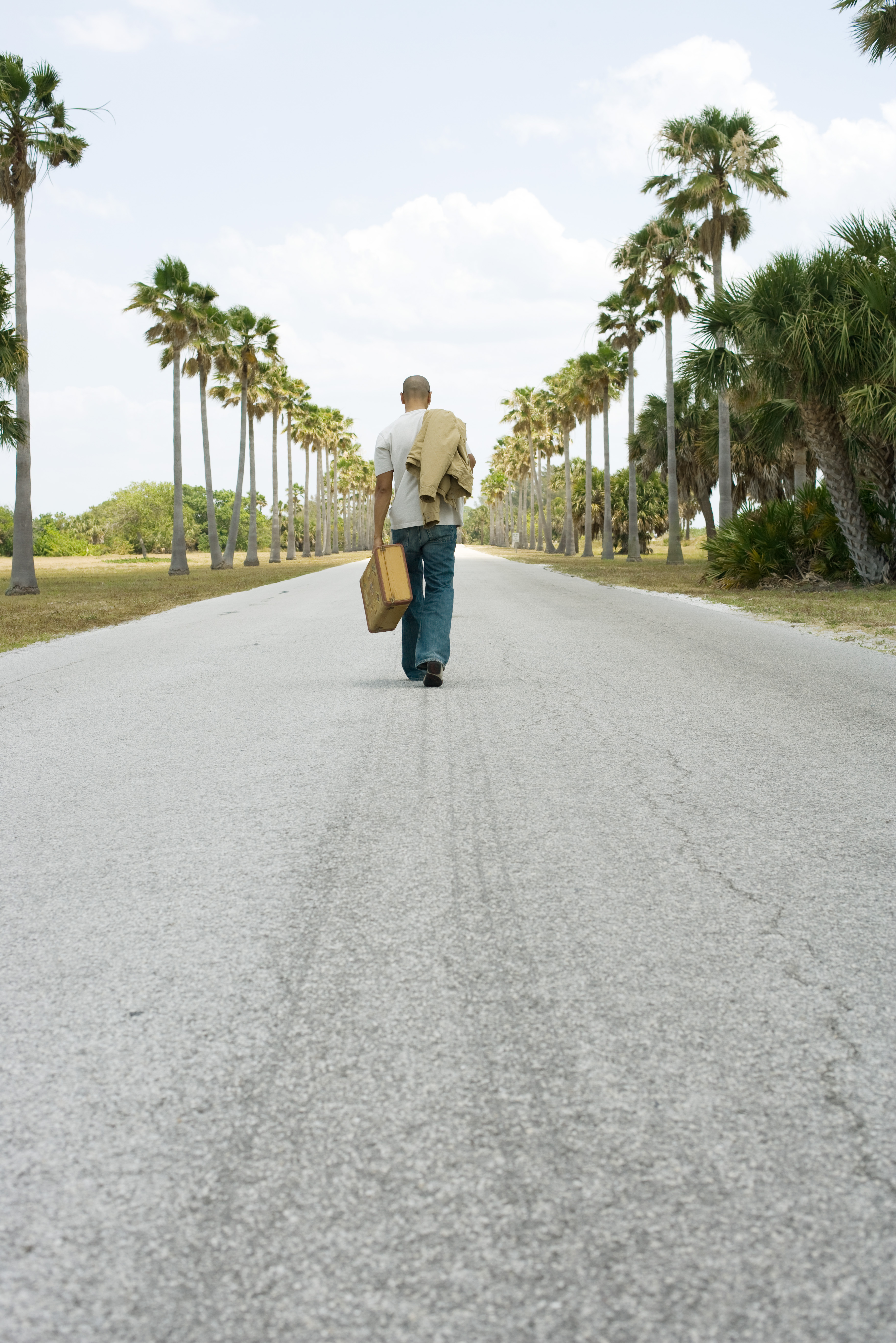 Man walking in center of road, carrying suitcase and jacket, rear view