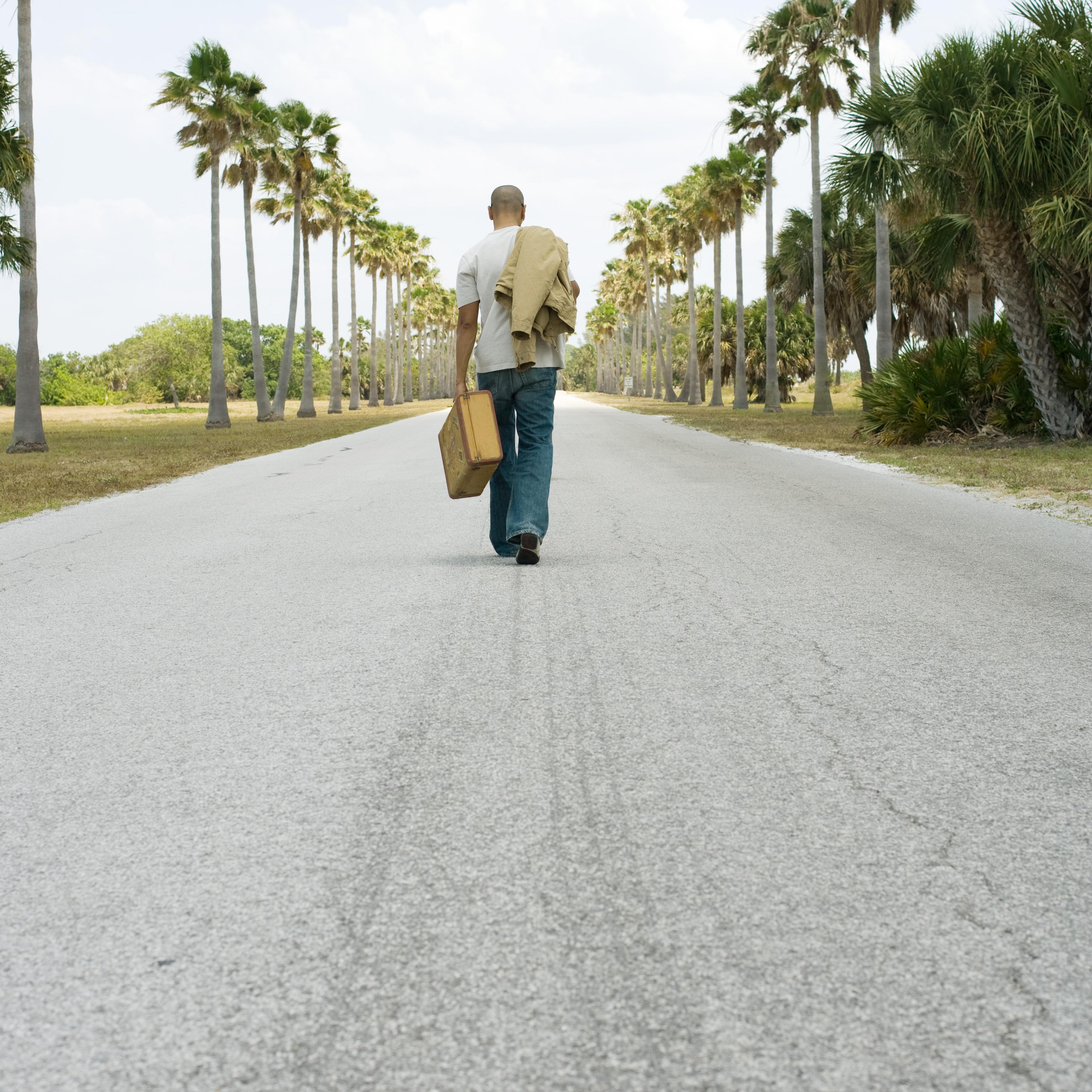 Man walking in center of road, carrying suitcase and jacket, rear view