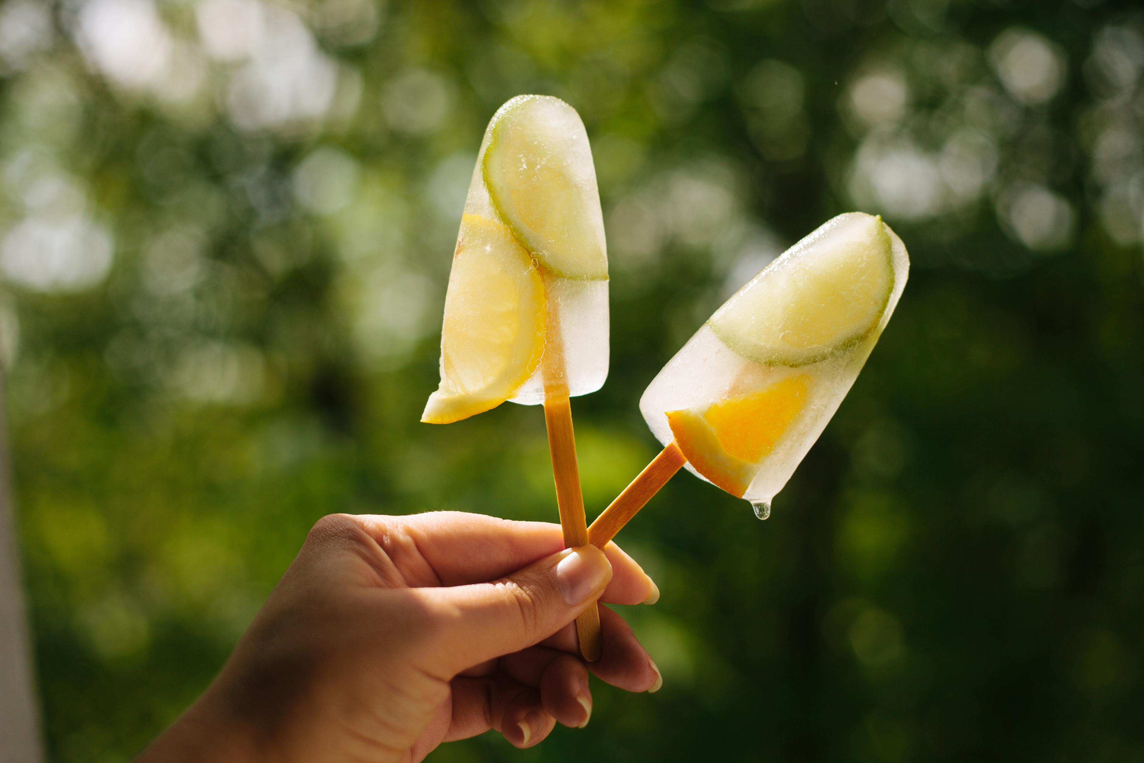 Two popsicles with lime and orange close-up in female hand against green background in bright sun light.