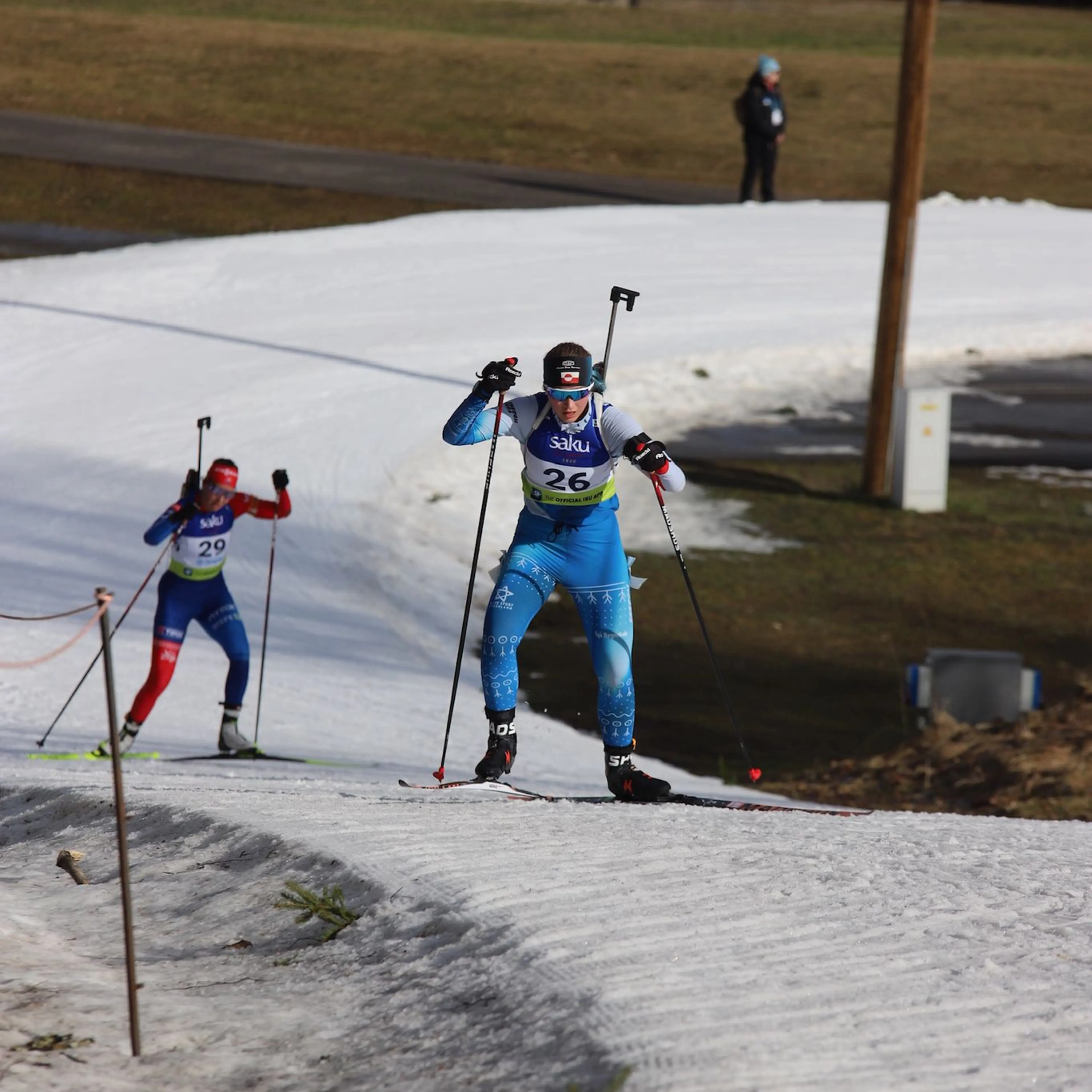 Two biathletes race on a narrow strip of snow with grass and mud on either side.