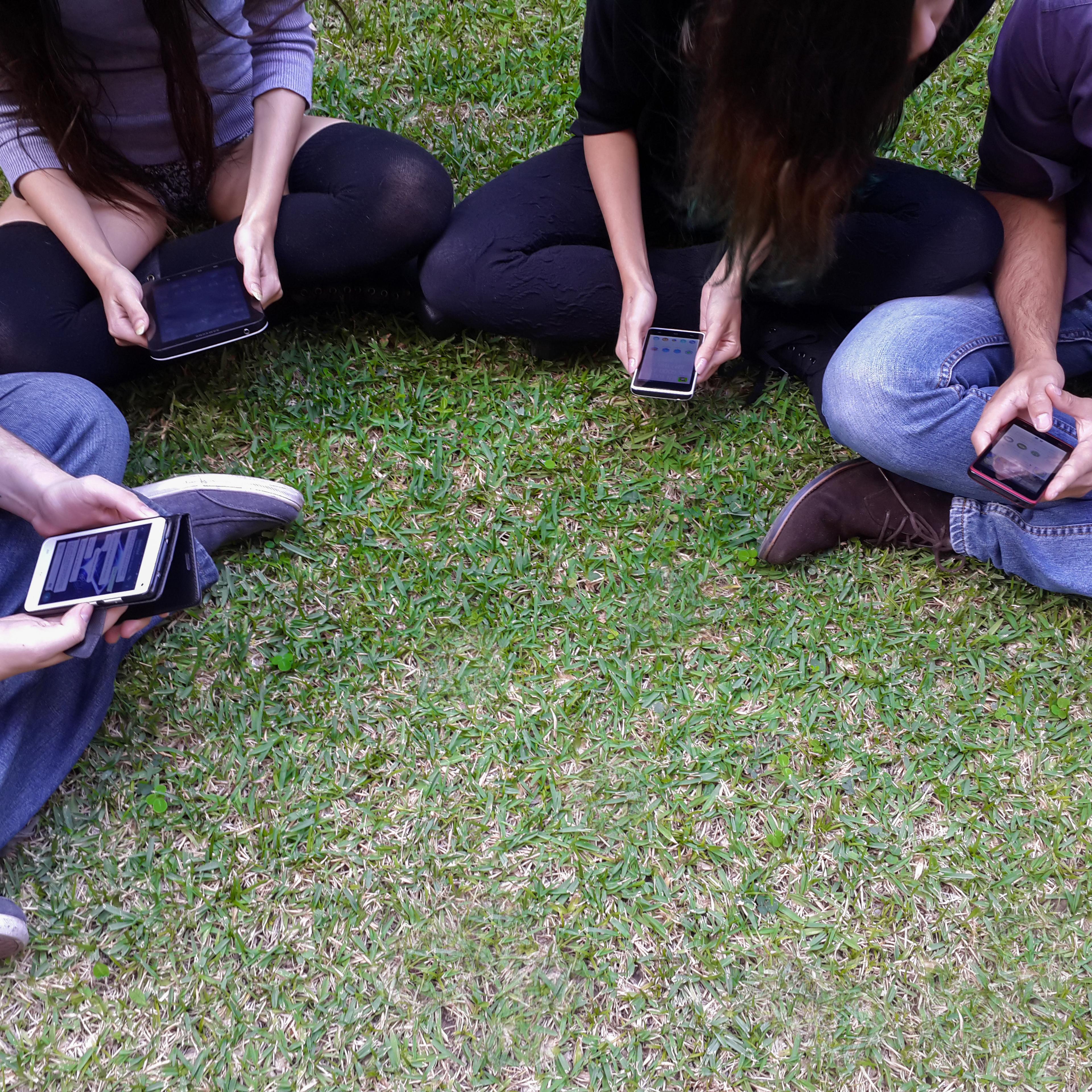 Millennials sit in a circle on grass looking at their phones.