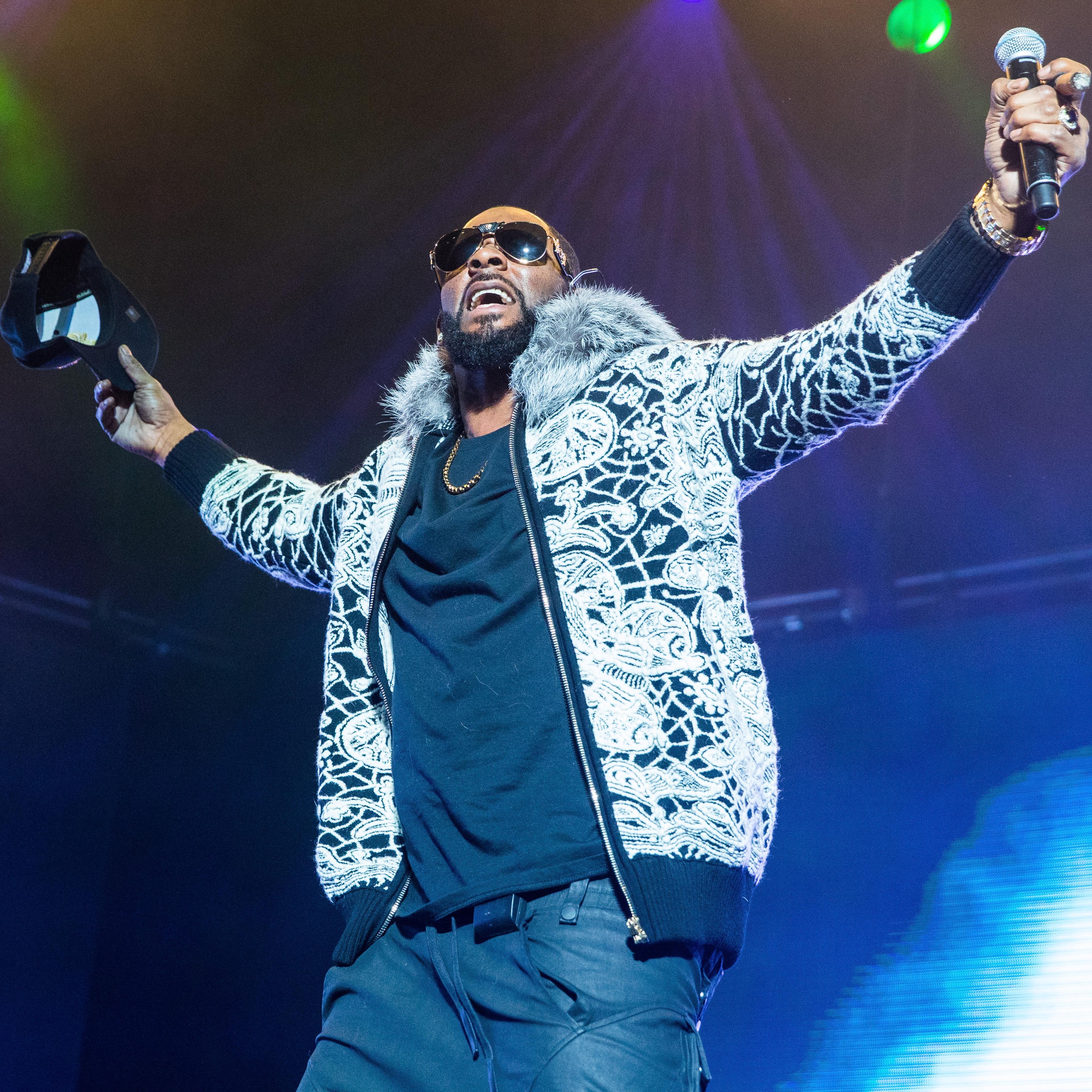 R. Kelly wears a fuzzy jacket and raises his arms to the side during concert
