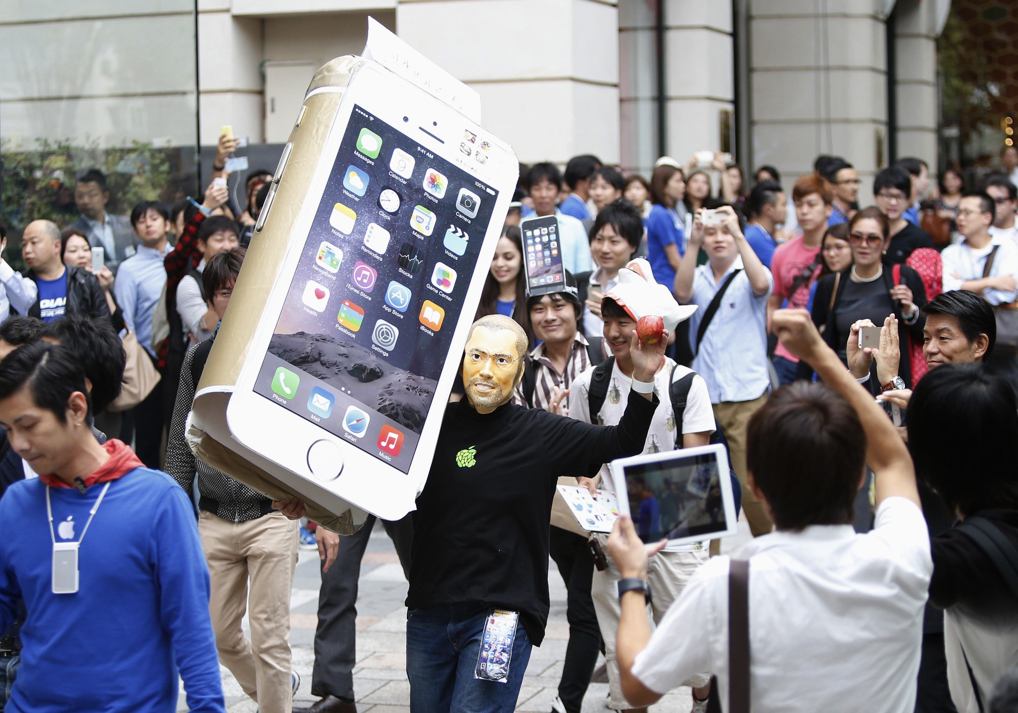 A man wearingaA man wearing a mask depicting Apple's co-founder Steve Jobs holds up a cardboard cut-out of Apple's new iPhone 6, as he walks into the Apple Store in Tokyo on Sept. 18, 2014. a mask depicting Steve Jobs holds up a cardboard cut-out of Appl