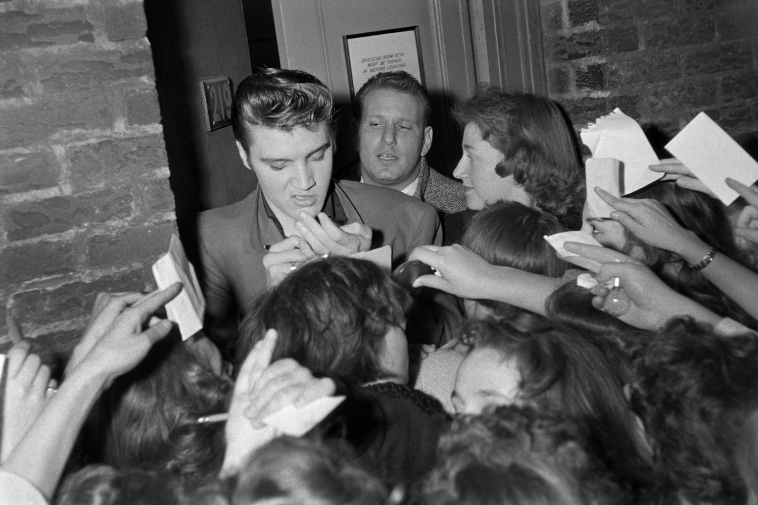 March 17, 1956. Studio 50 New York. This was the crowd after the Dorsey Brothers 'Stage Show.'  I just like the snarl. There were about a hundred well wishers at the backstage entrance and fans asking for autographs. The girl on the right was helping with slips of paper — she was so happy to be close to him. And this guy was concerned with keeping Elvis warm. 
