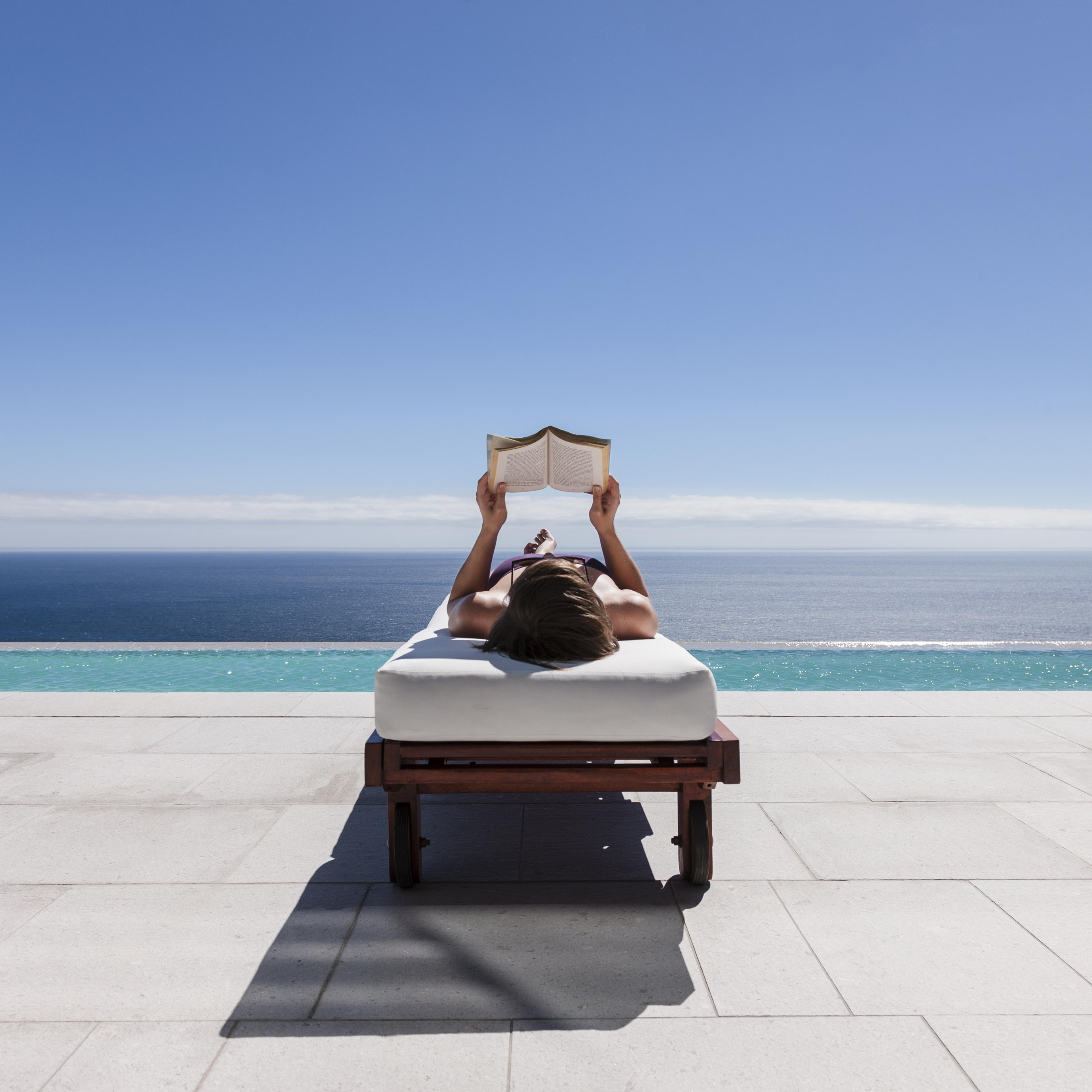 Woman reading on lounge chair at poolside overlooking ocean