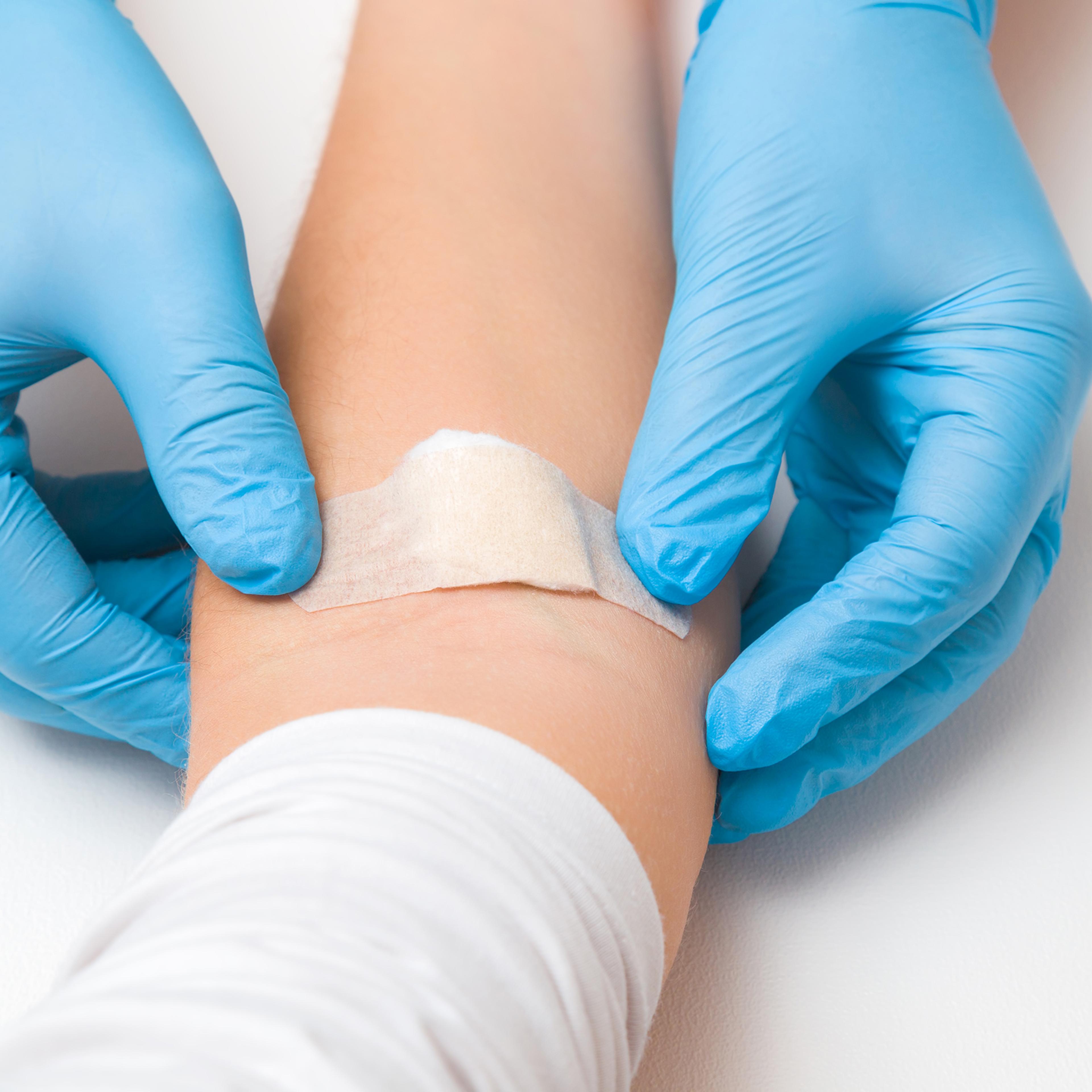 Doctor in blue rubber protective gloves putting an adhesive bandage on young woman's arm vein after blood test or injection of vaccine. First aid. Medical, pharmacy and healthcare concept. Closeup.