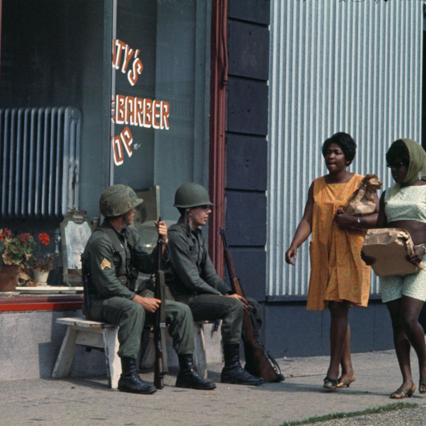 Two women walk past National Guardsmen seated on a bench outside a shop during rioting in Detroit, Mich., July 27 1967.