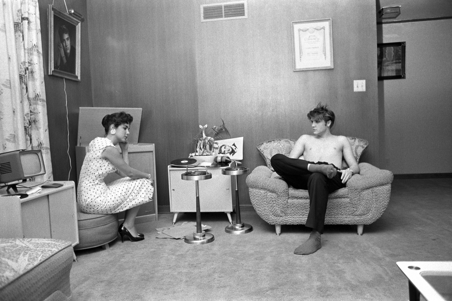 July 4, 1956. 1034 Auburn Drive, Memphis. With Barbara Hearn listening to playback. The shot of Elvis on the wall is his first publicity shot (the gold frame with a light over the top.) And his mother liked it so much that she went back to the photographer and had it hand painted in color on canvas…looked like a museum painting!  Here, Elvis is testing the three songs that he recorded a few days earlier. He had gotten a quick acetate cut, which would play for about 50 takes before it disintegrated. He tried to dance with her, but he was all sweated up, and she said "after you're showered up." He wanted to know if the music was danceable — she said no.