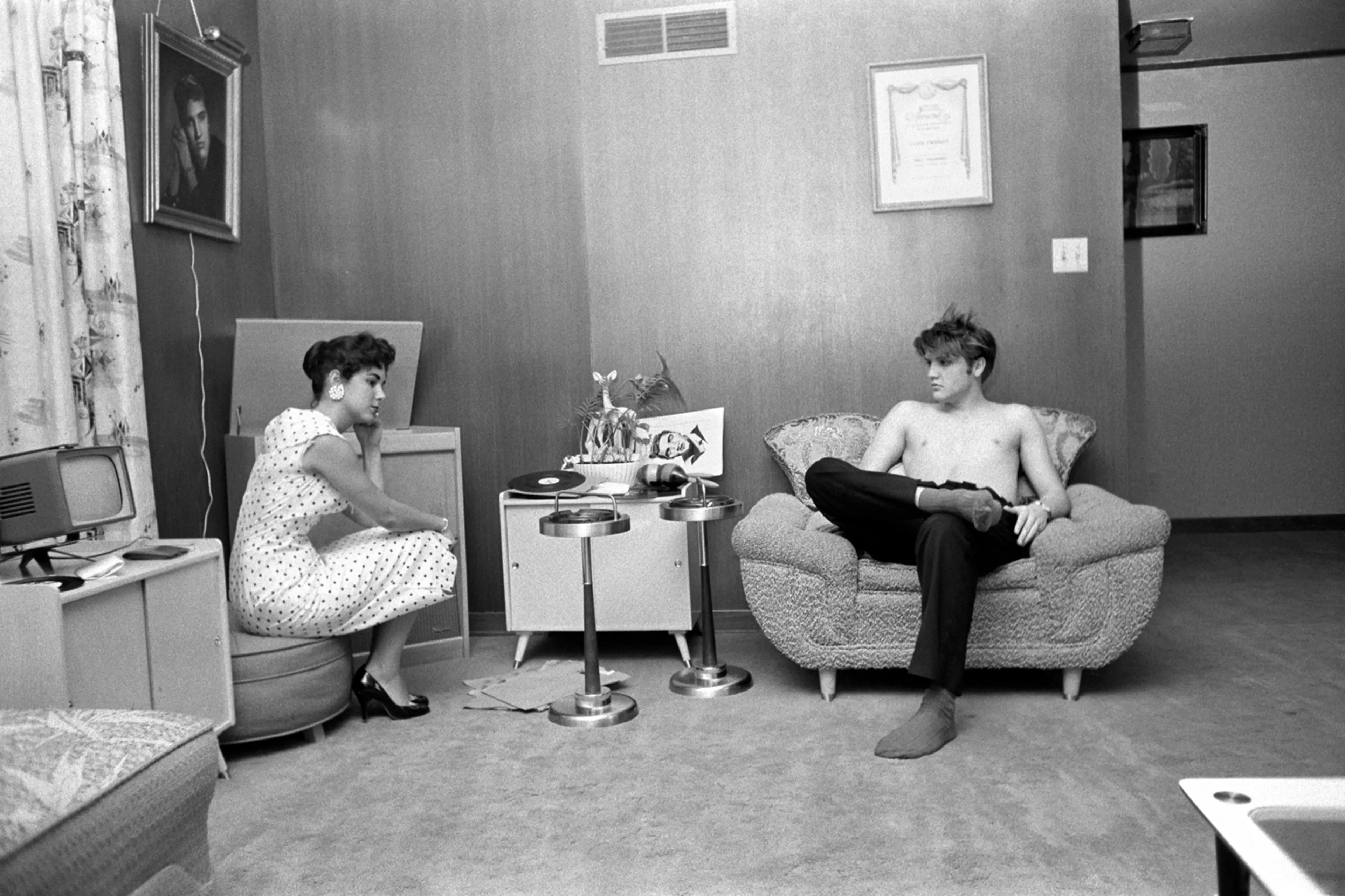 July 4, 1956. 1034 Auburn Drive, Memphis. With Barbara Hearn listening to playback. The shot of Elvis on the wall is his first publicity shot (the gold frame with a light over the top.) And his mother liked it so much that she went back to the photographer and had it hand painted in color on canvas…looked like a museum painting! Here, Elvis is testing the three songs that he recorded a few days earlier. He had gotten a quick acetate cut, which would play for about 50 takes before it disintegrated. He tried to dance with her, but he was all sweated up, and she said "after you're showered up." He wanted to know if the music was danceable — she said no.
