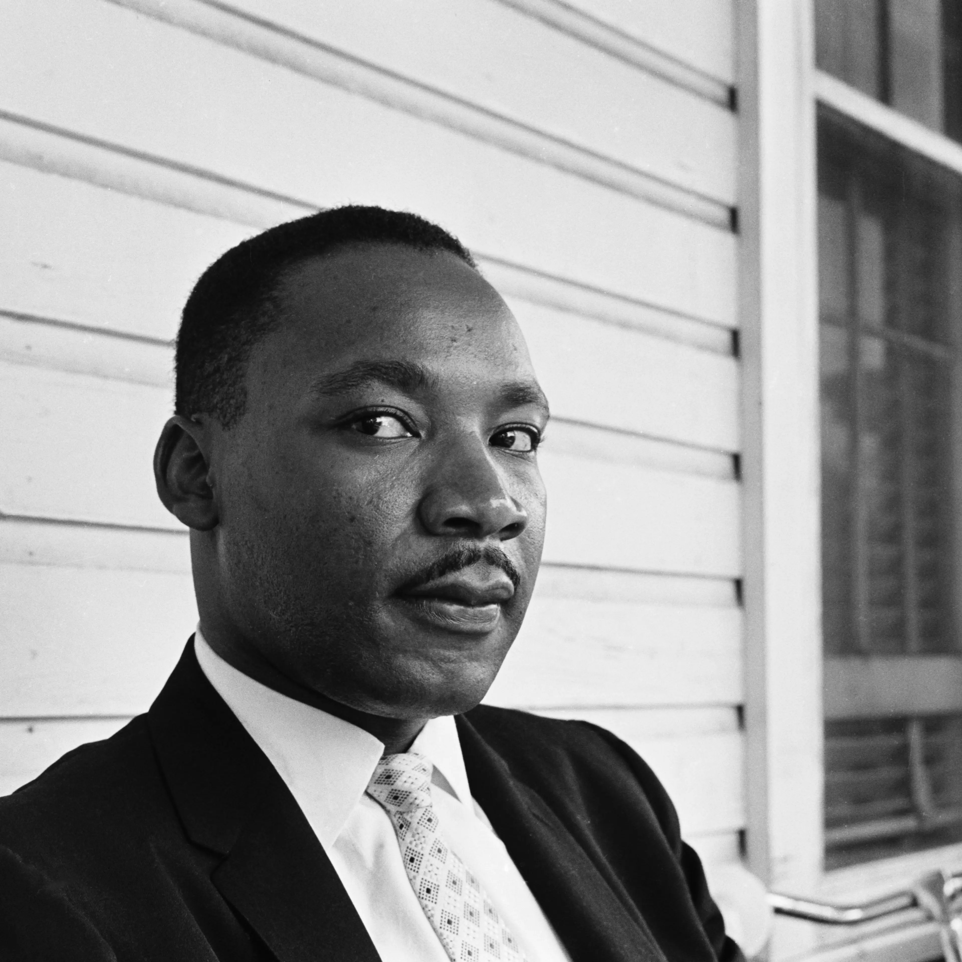 In this black and white portrait of The Rev. Dr. Martin Luther King, Jr., he wears a suit and tie and looks pensively at the camera.