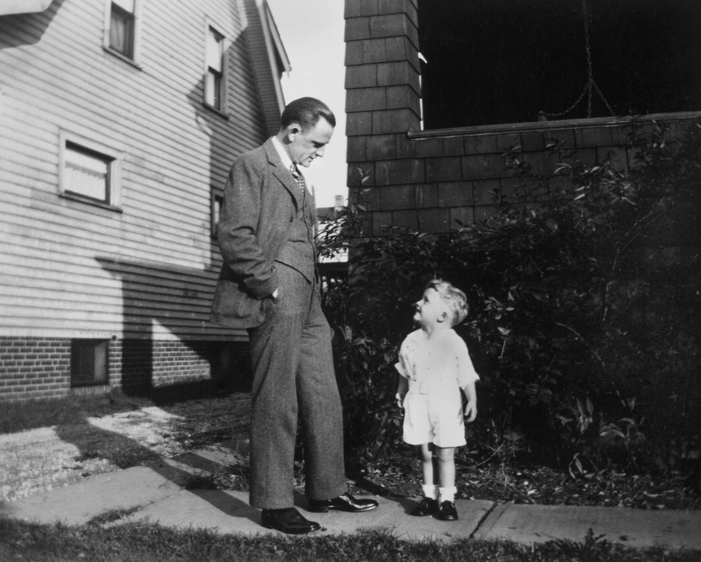 Father and son look at each other on sidewalk, ca. 1930.