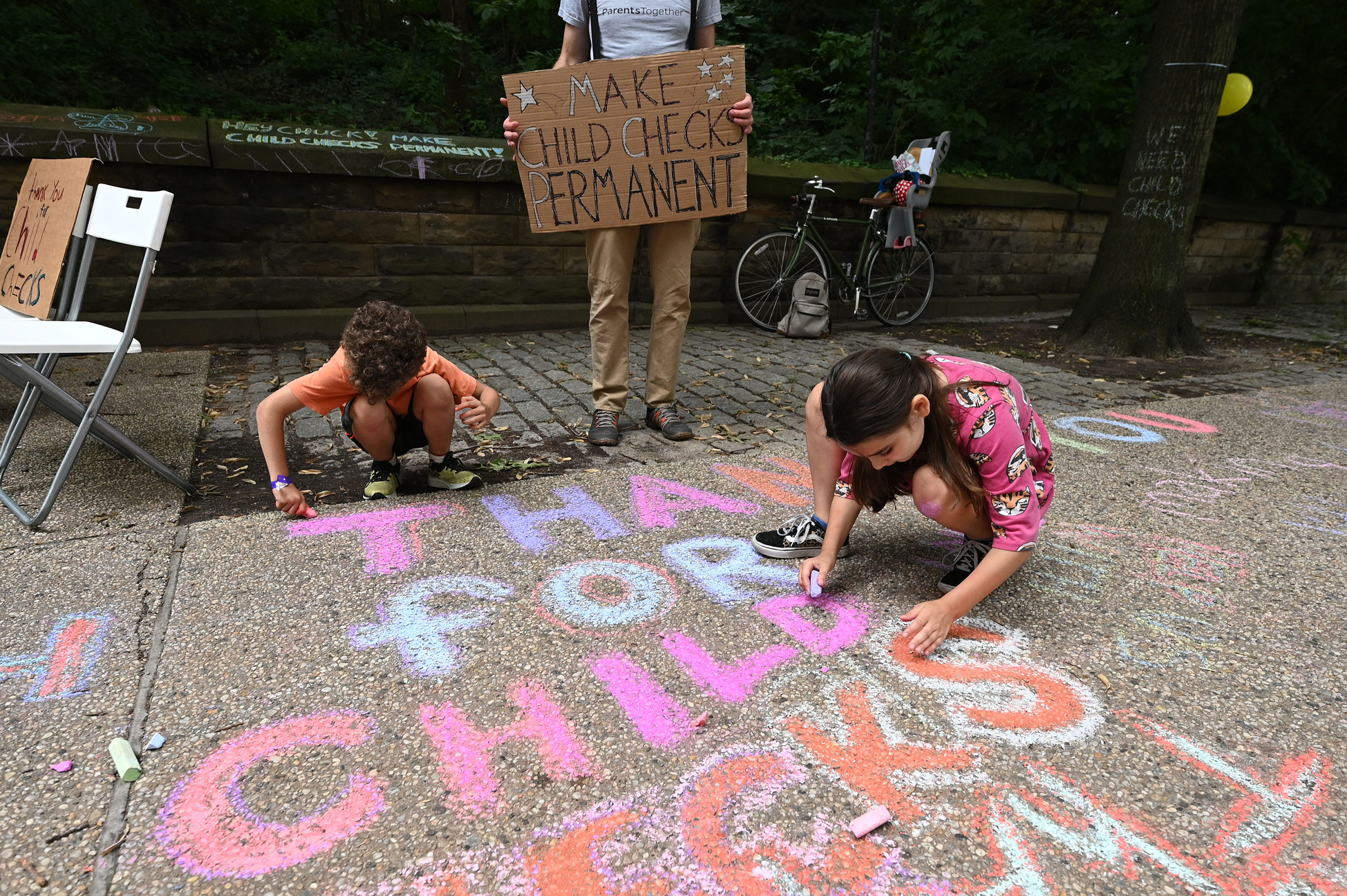 Justin Ruben, Rime Leonard and Edie Abrams-Pradt draw with chalk to celebrate new monthly Child Tax Credit payments and urge congress to make them permanent outside Senator Schumer's home on July 12, 2021 in Brooklyn, New York.