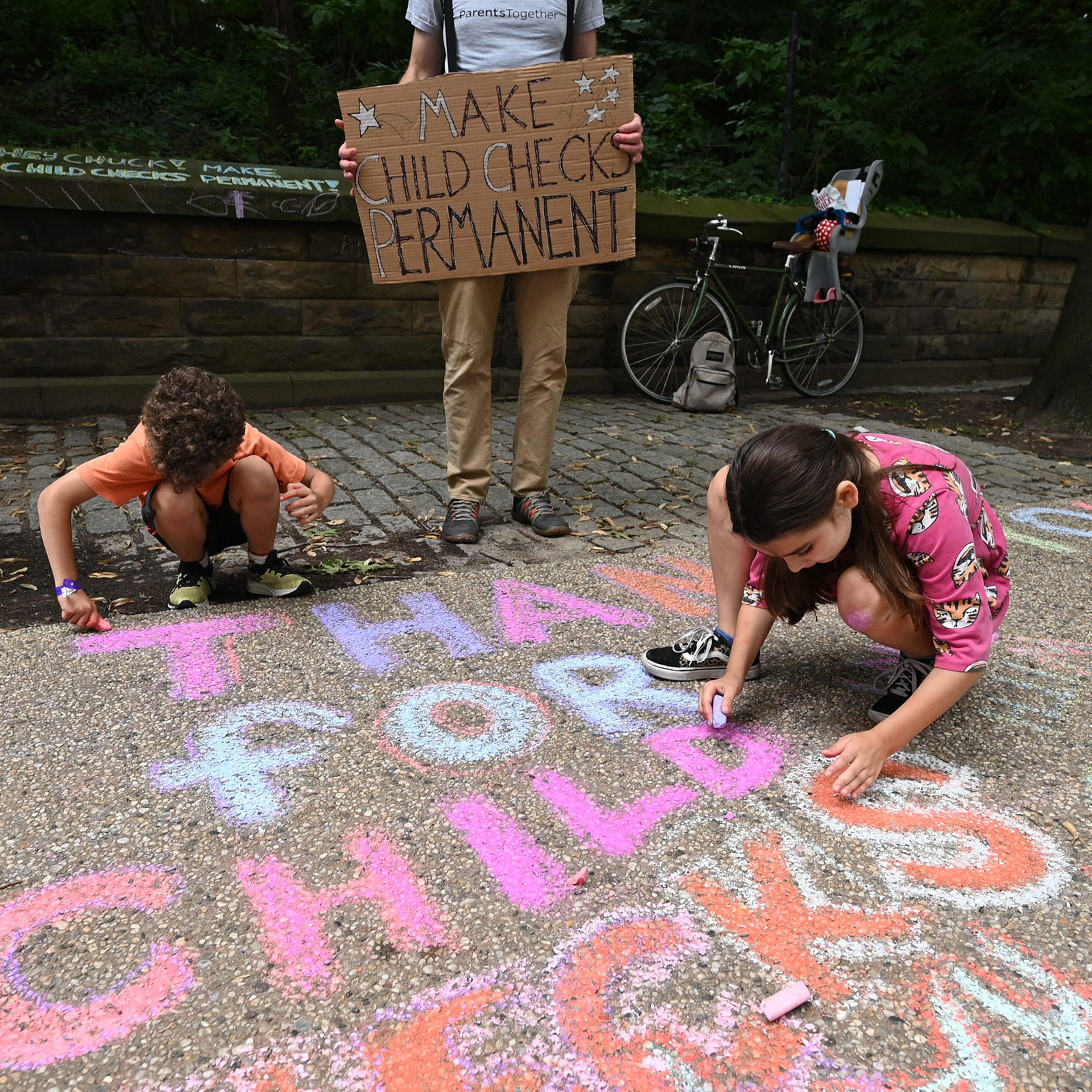 Justin Ruben, Rime Leonard and Edie Abrams-Pradt draw with chalk to celebrate new monthly Child Tax Credit payments and urge congress to make them permanent outside Senator Schumer's home on July 12, 2021 in Brooklyn, New York.