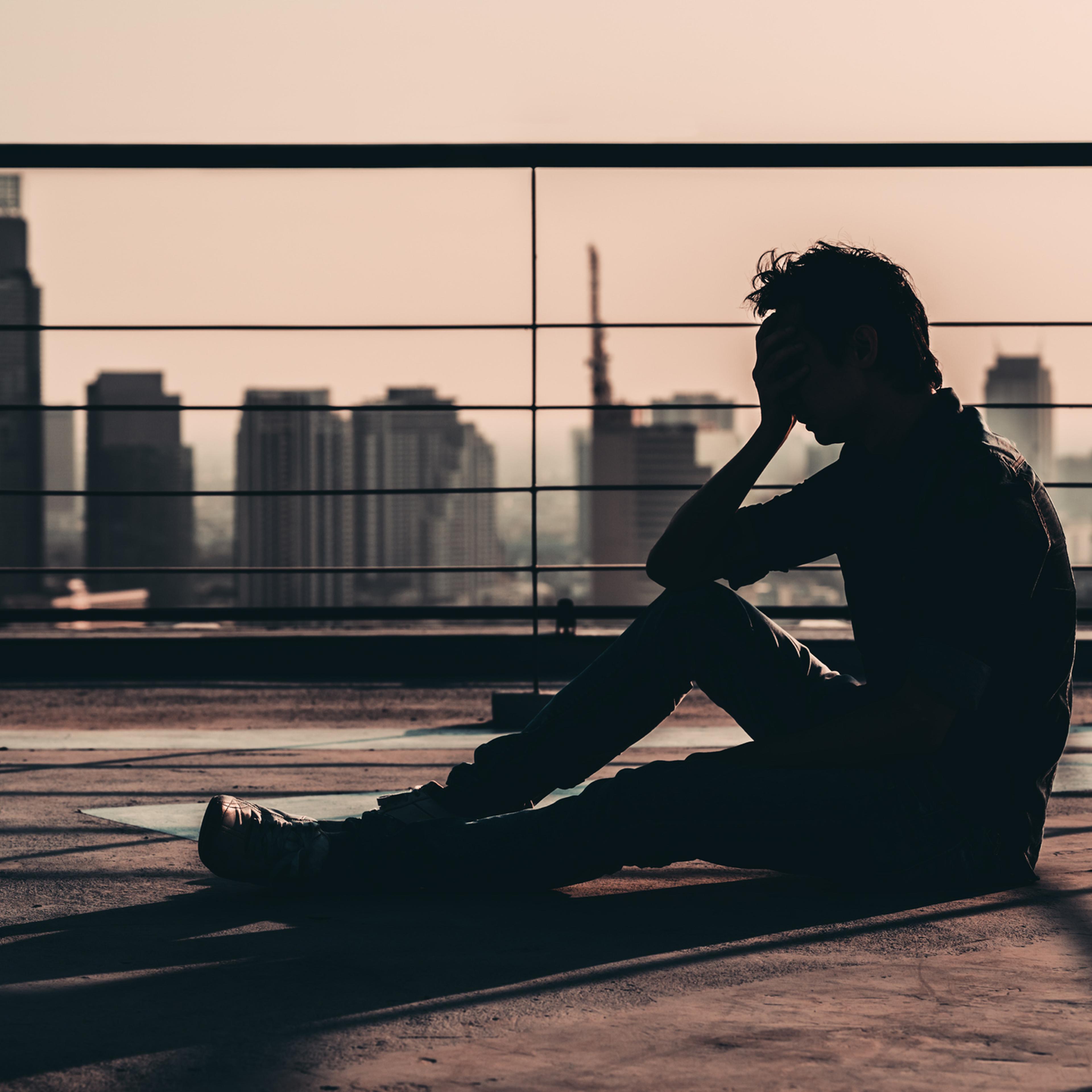 Silhouette of sad depressed Asian man lost hope and cry, sit on building rooftop at sunset, dark mood tone. Concept of major depressive disorder, friend zone, unemployment, stress emotion or paranoid
