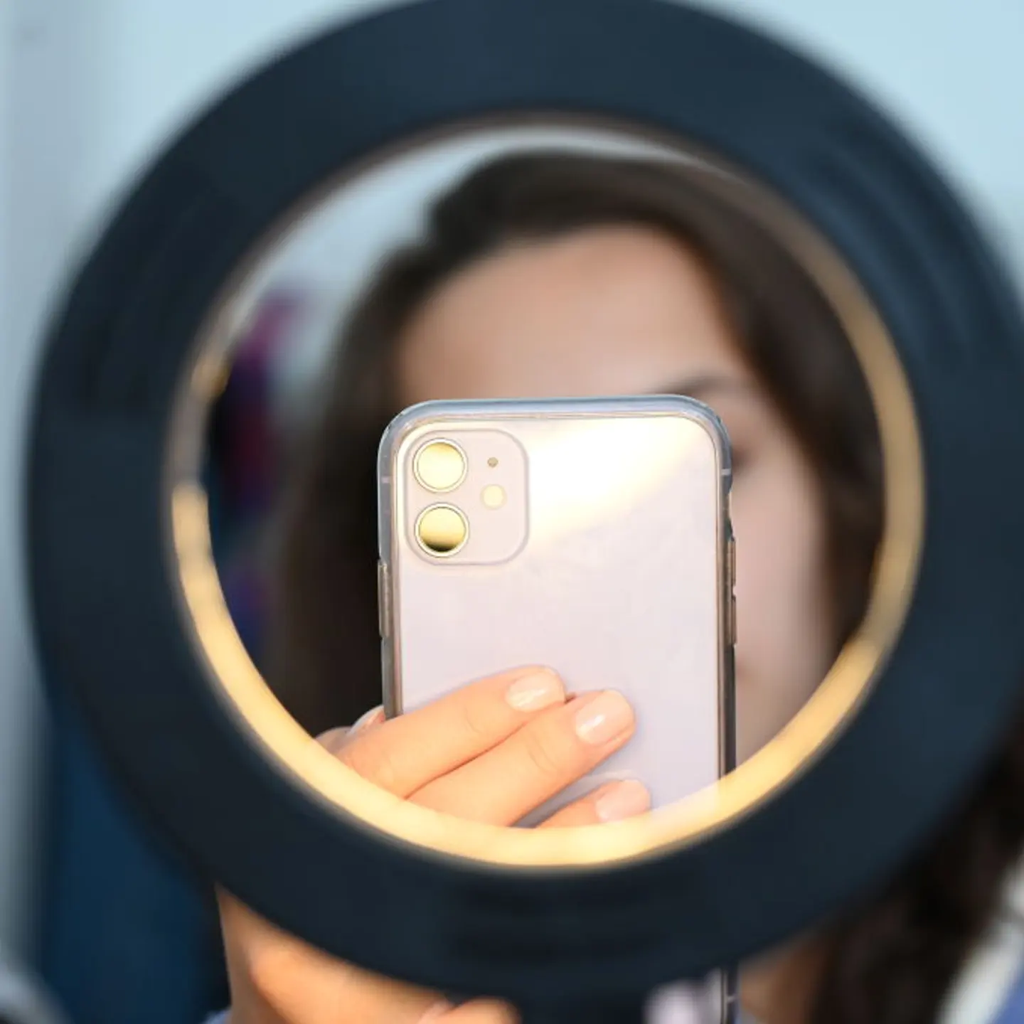 A woman sits behind a ring light and takes pictures of herself with her phone camera.