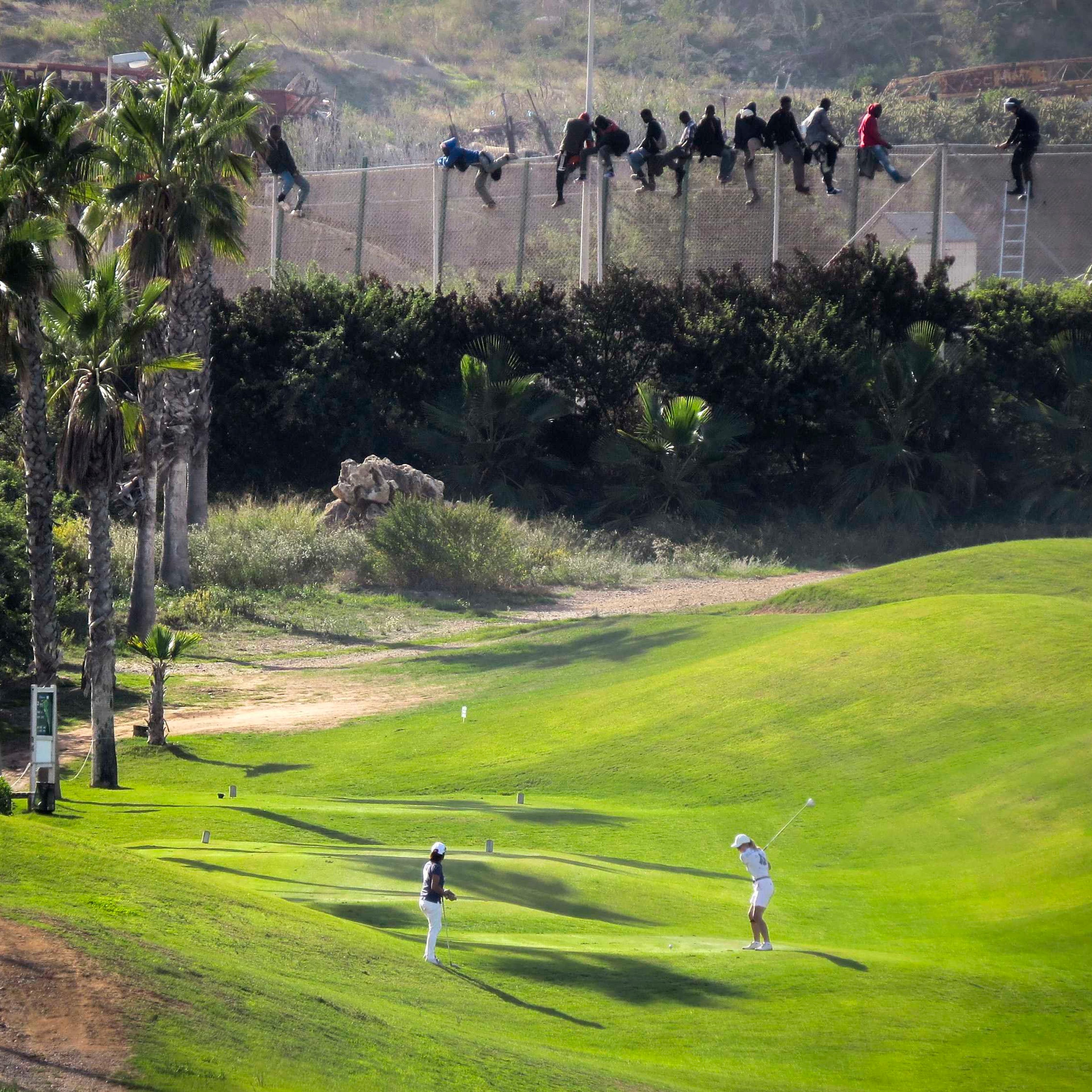 A golfer hits a tee shot as African migrants sit atop a border fence during an attempt to cross into Spanish territories between Morocco and Spain's north African enclave of Melilla