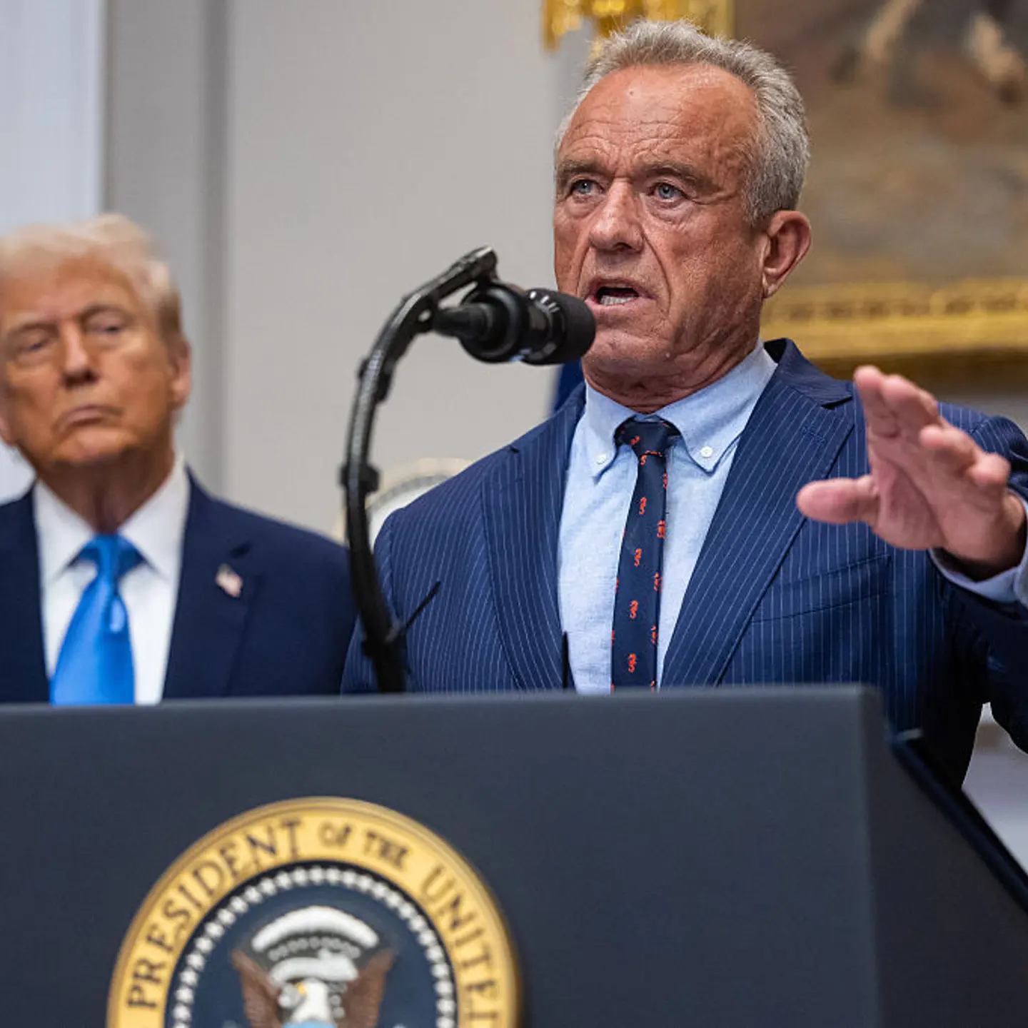 Robert F. Kennedy Jr. stands in front of a podium speaking. Trump is in the background.