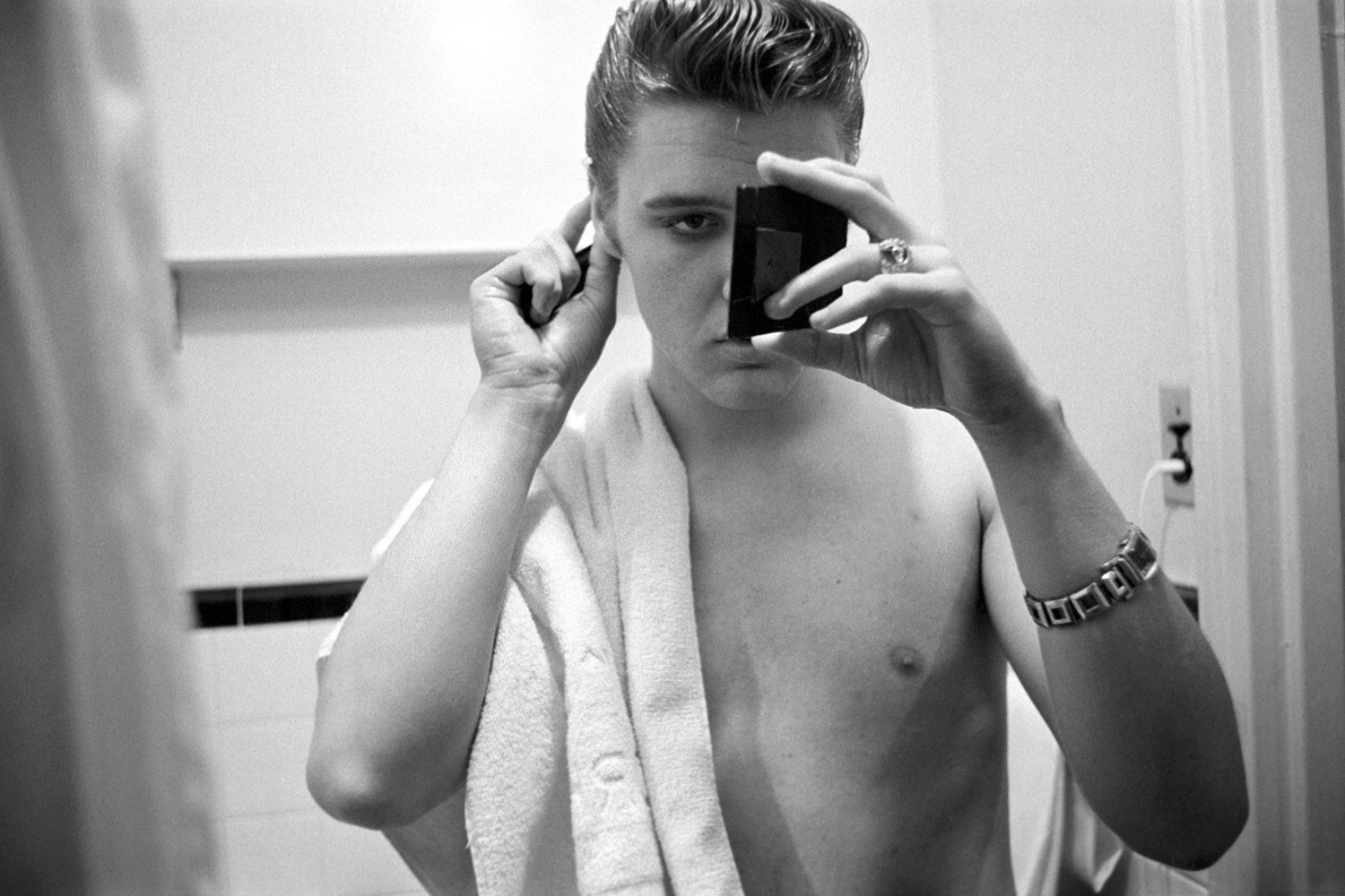 March 17, 1956. The Warwick New York. Elvis in his hotel bathroom an hour before returning to perform on 'Stage Show.' I love this one…he’s got full concentration. Elvis had just finished showering — he's looking into a small ladies hand mirror to see what's going on with the back of his hair, in a bigger mirror. He's using Vaseline hair tonic, not "bear grease." 