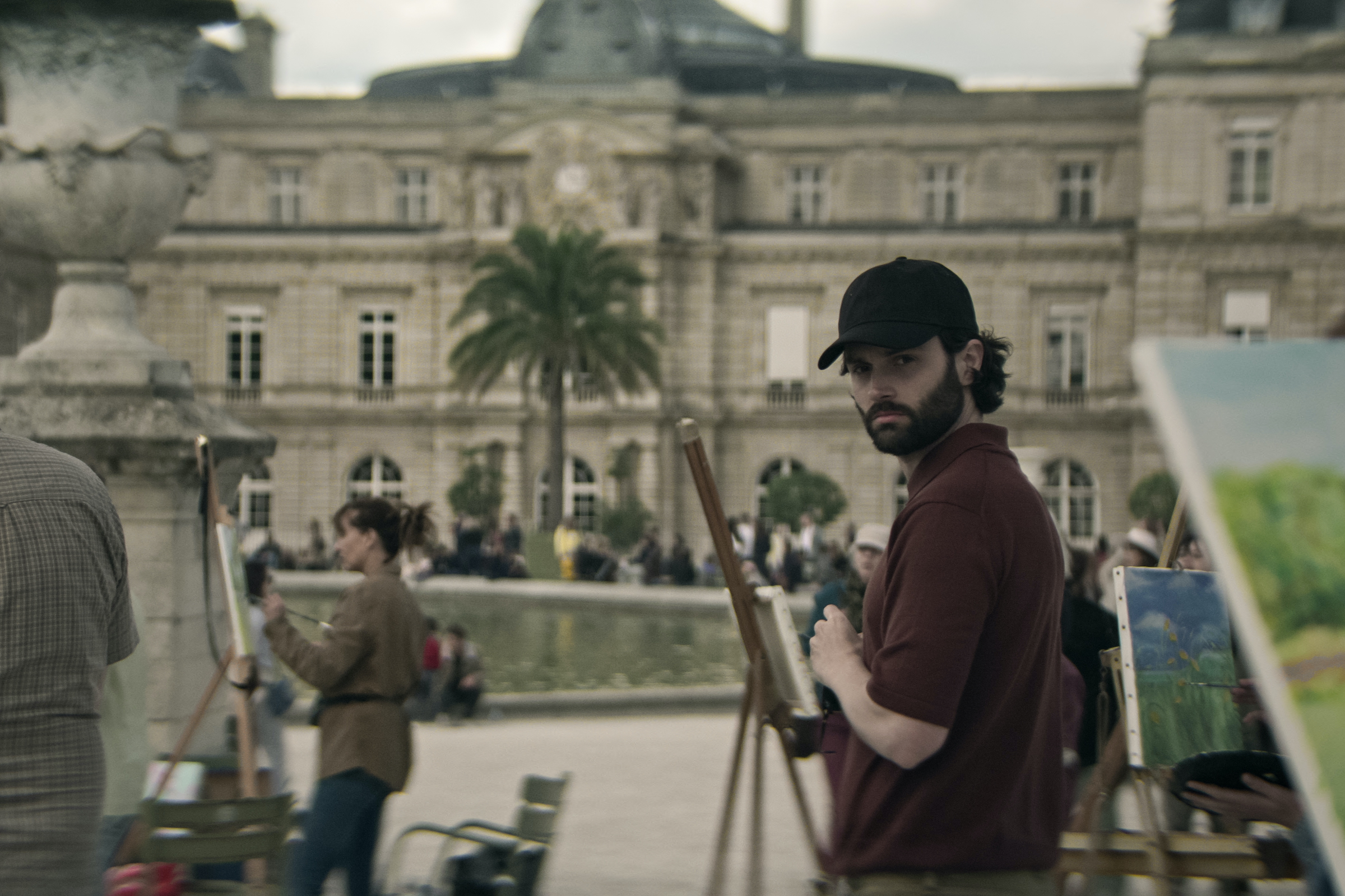 Penn Badgley turns and looks directly into the camera in a brown polo and black baseball cap.
