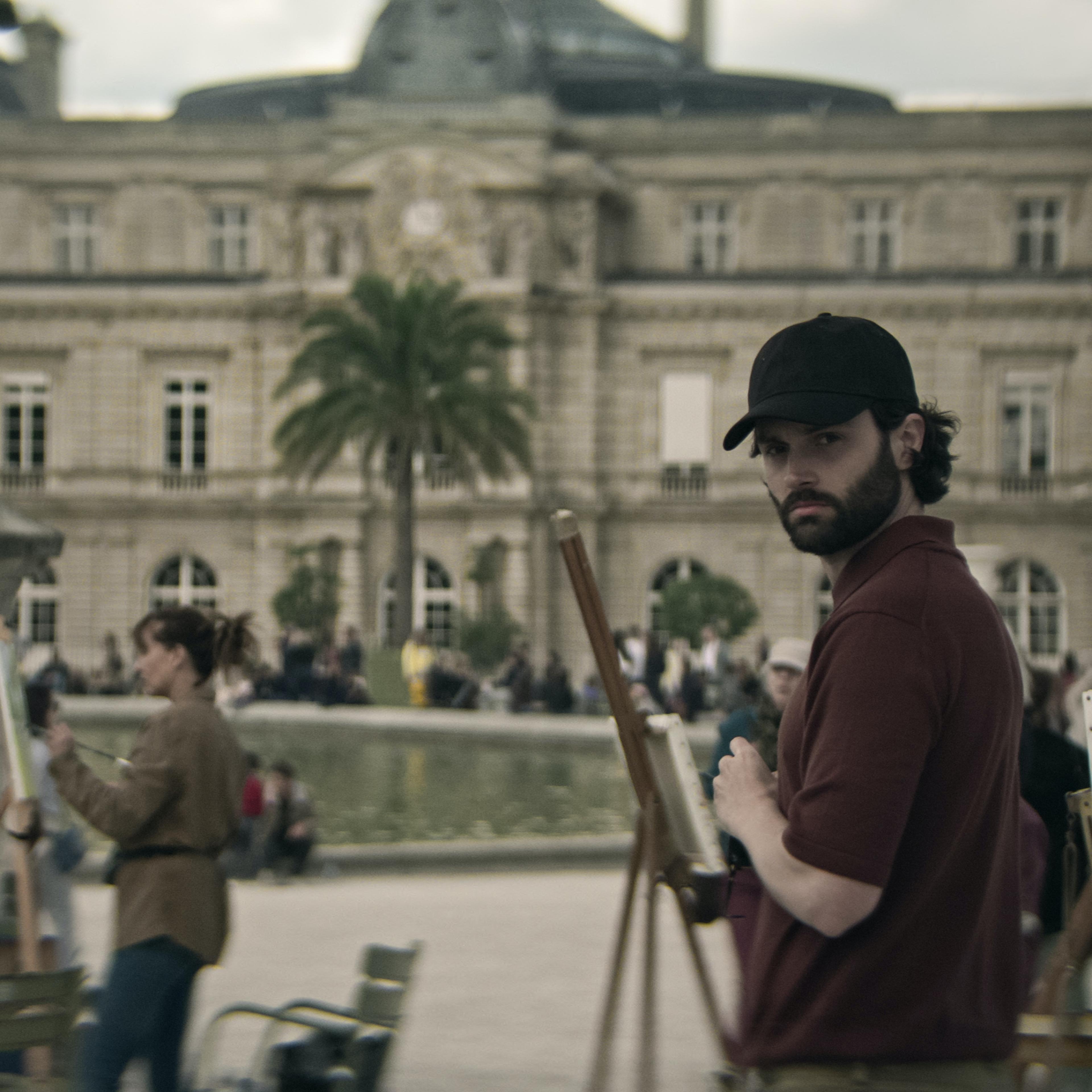 Penn Badgley turns and looks directly into the camera in a brown polo and black baseball cap.