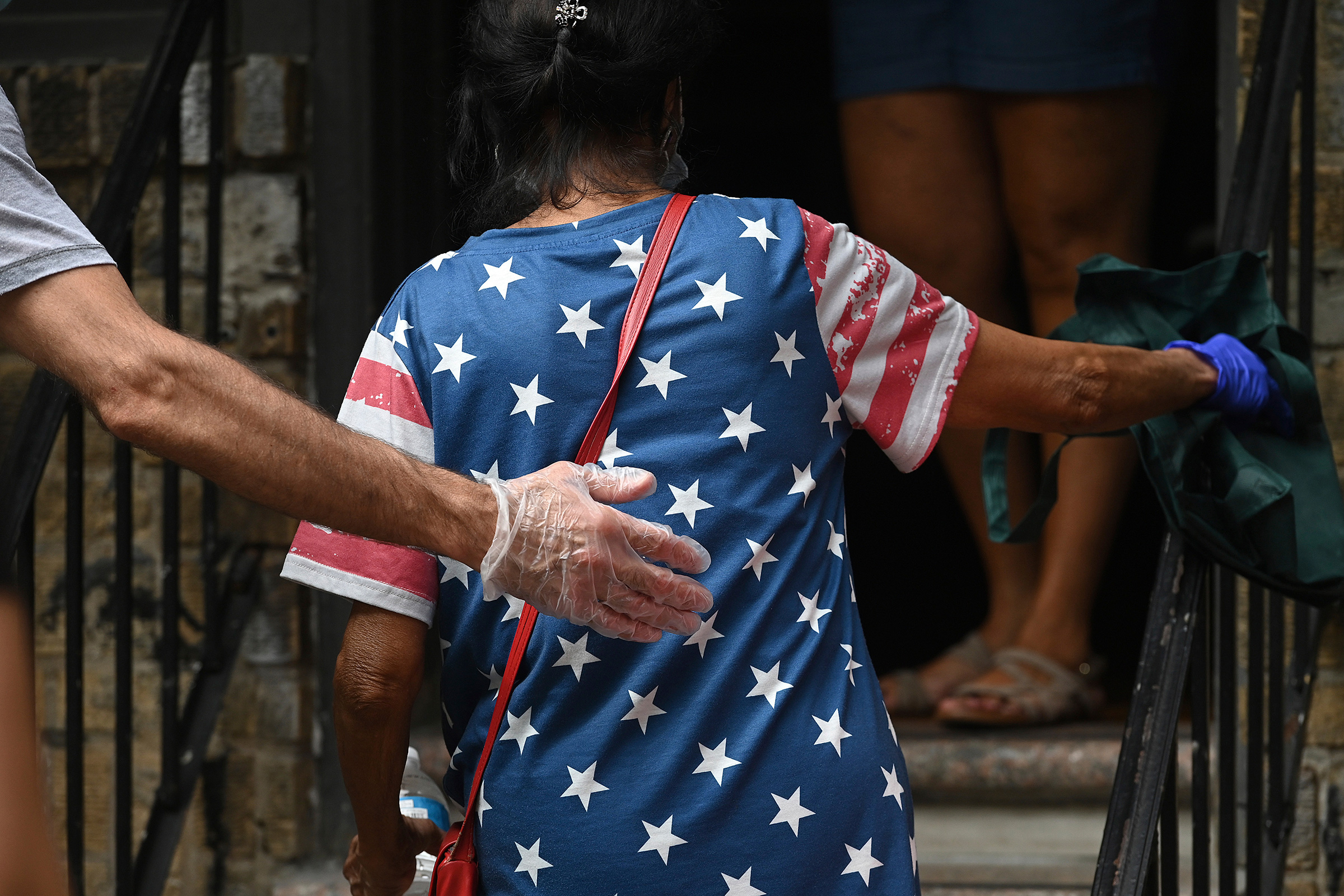 With an outstretched gloved hand, a volunteer helps a woman up the steps of the Mosaic Community Center Food Pantry to receive donated food in Queens on July 31