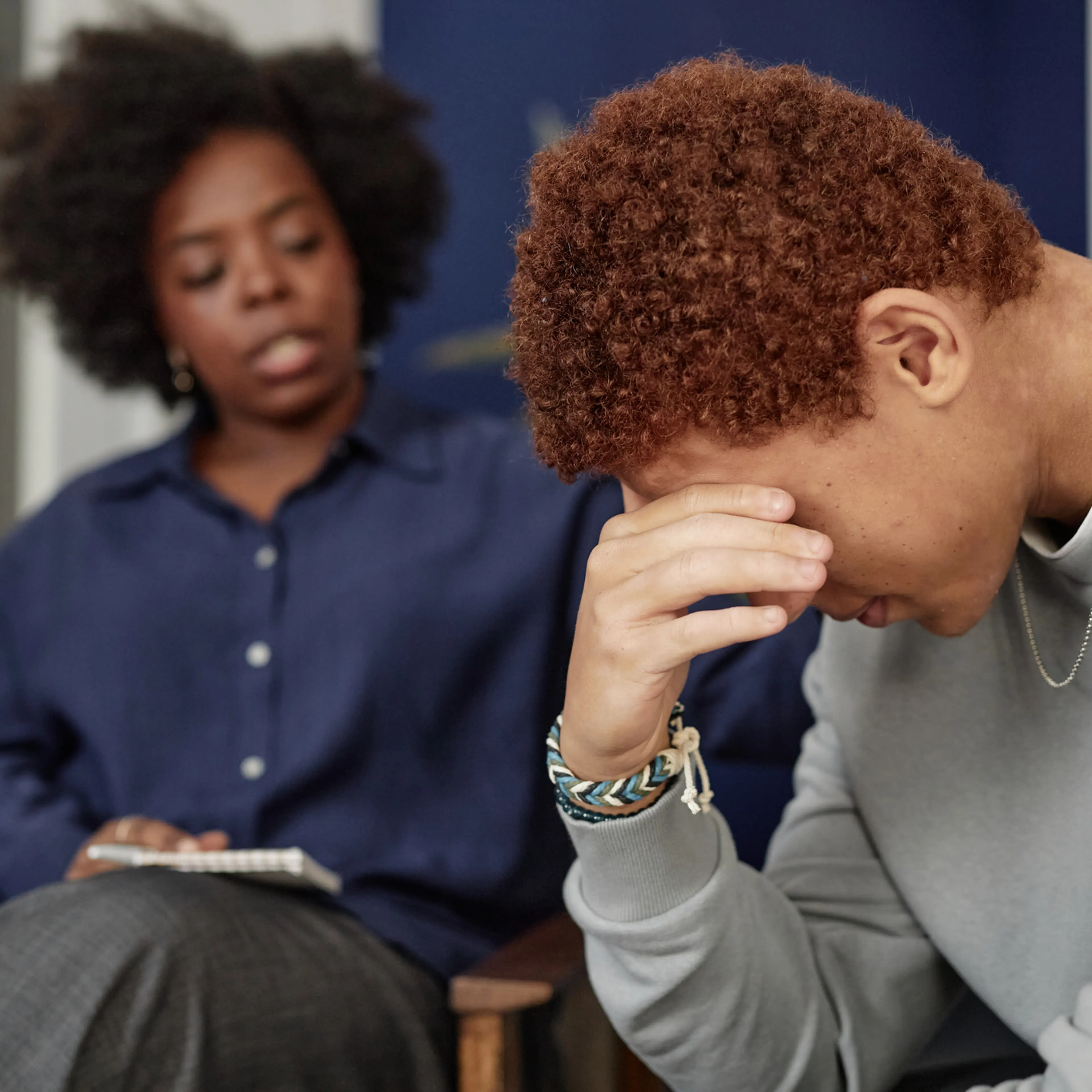 Side view closeup of Black teenage boy crying during support group meeting with woman therapist comforting him in blurred background copy space