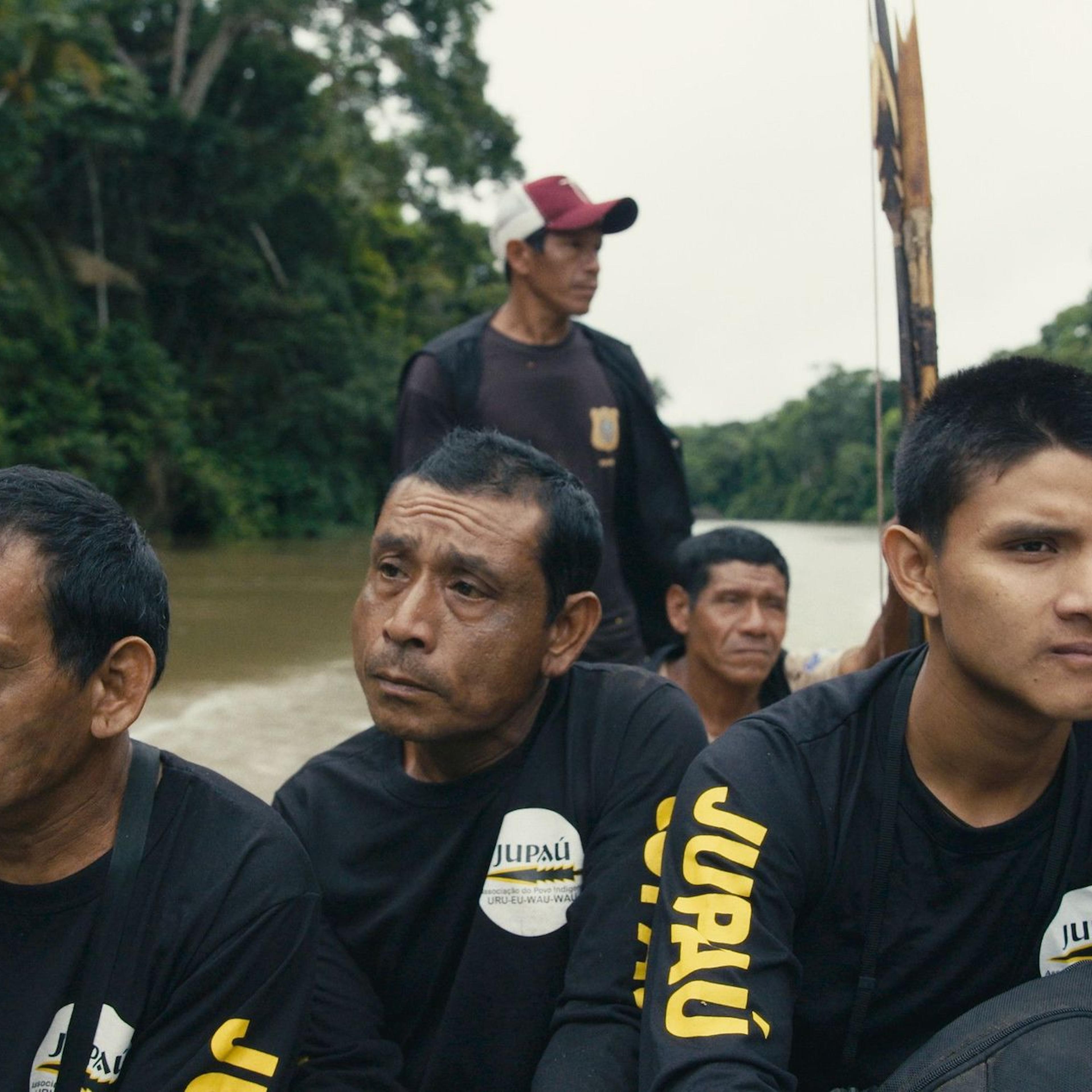Five Indigenous men sit in a boat racing toward the camera, all looking slightly away