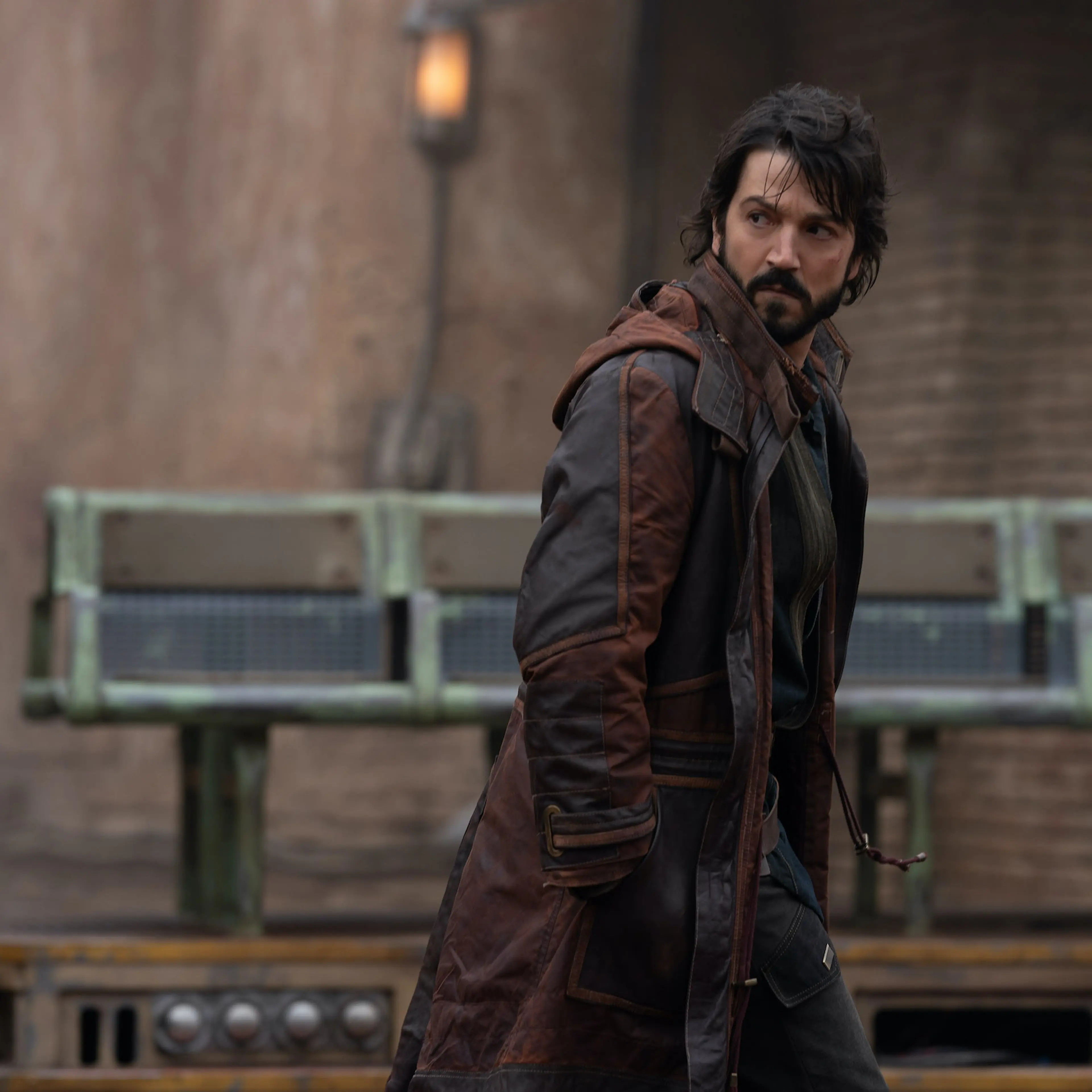 Diego Luna, dressed in a long brown coat, walks toward the right of the frame, looking behind him.