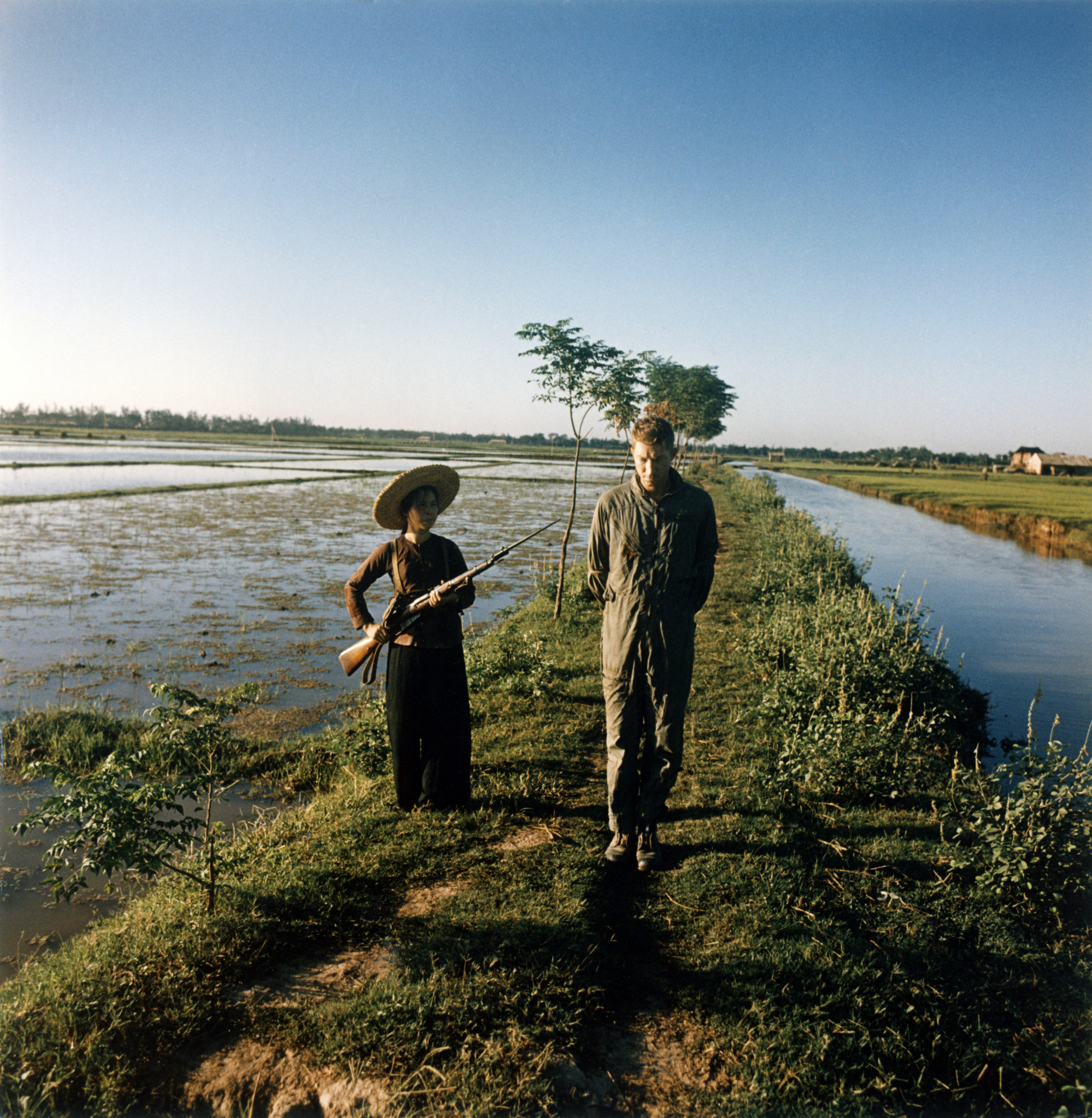 The captured US pilot major Pewey Waddell is being guarded by a militiawoman with a gun and a bayonet on a rice field - 1967