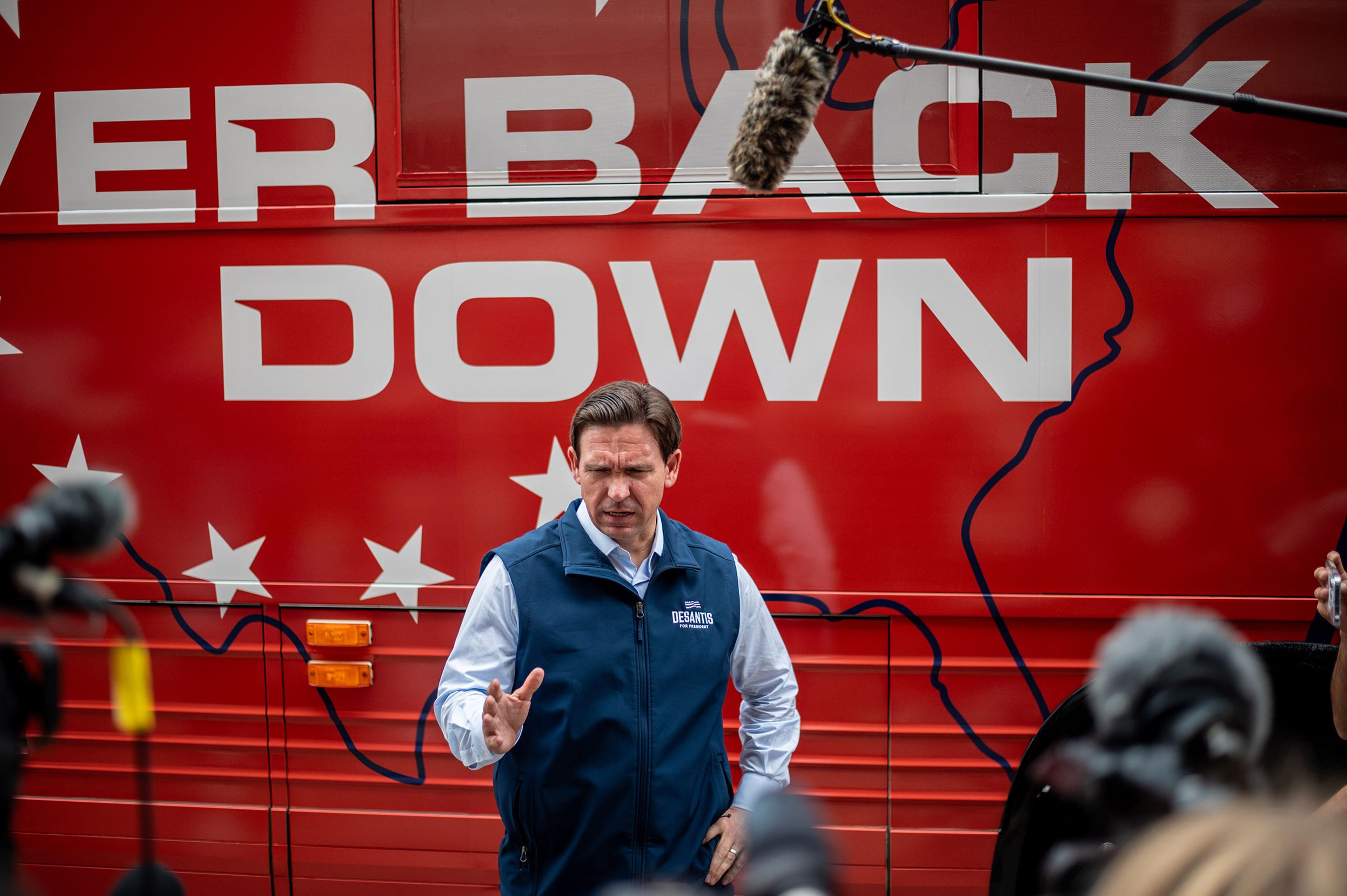 Ron DeSantis stands in front of his red tour bus that has the saying "never back down" on it as he gestures and speaks to members of the press