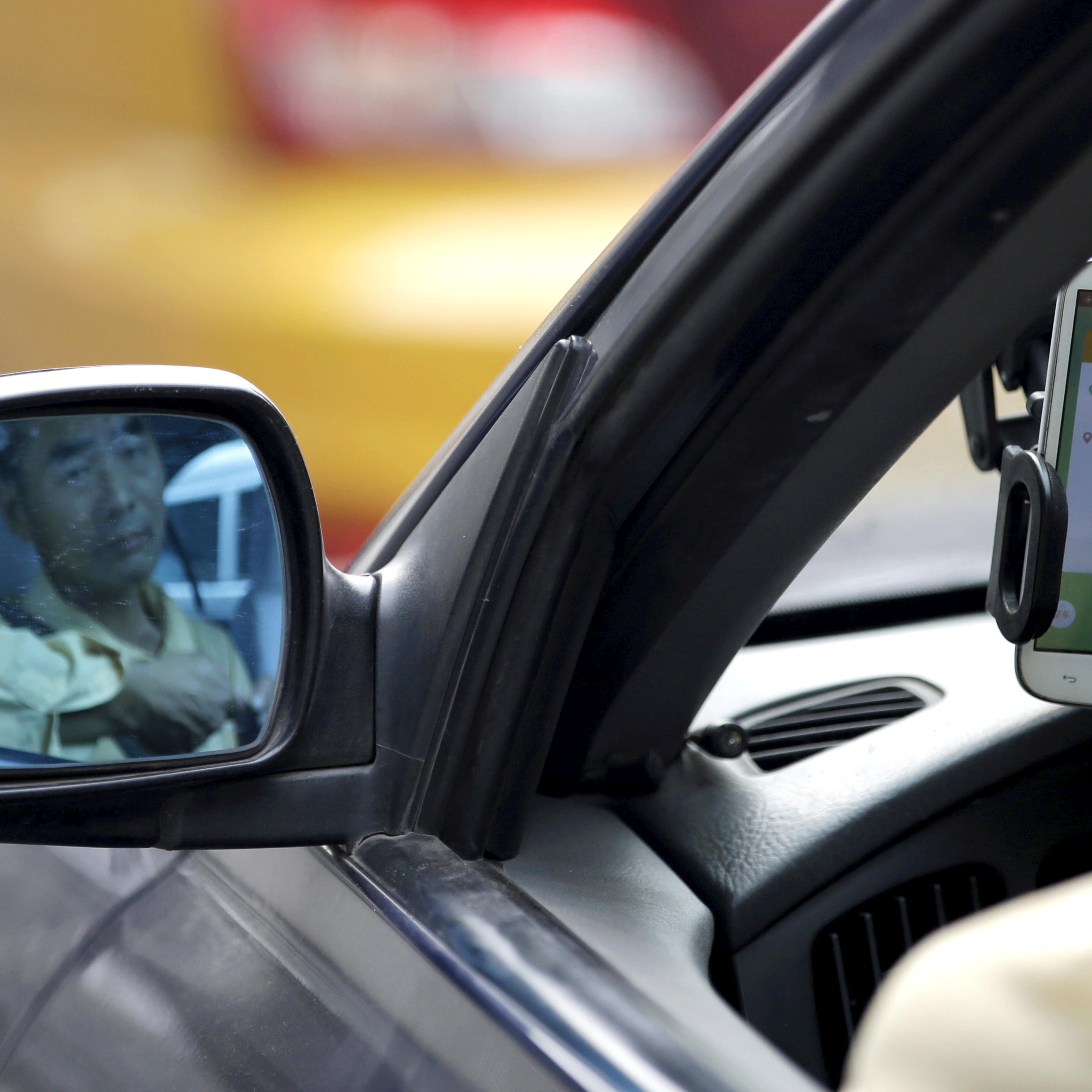 A taxi driver is reflected on a side mirror as he uses Didi Chuxing car-hailing application in Beijing